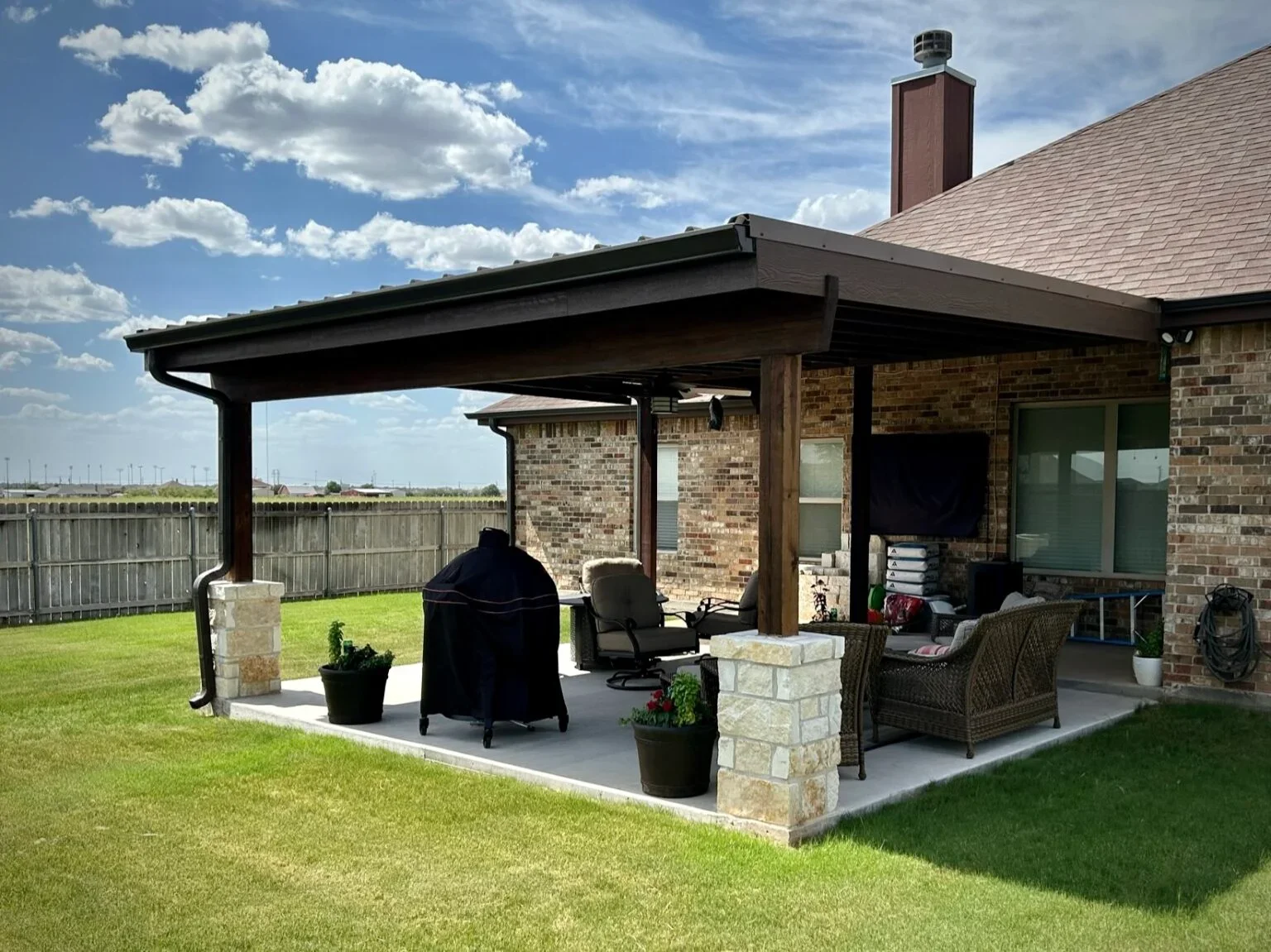 Backyard patio with a covered seating area, brick house, lawn, and a wooden fence under a partly cloudy sky.