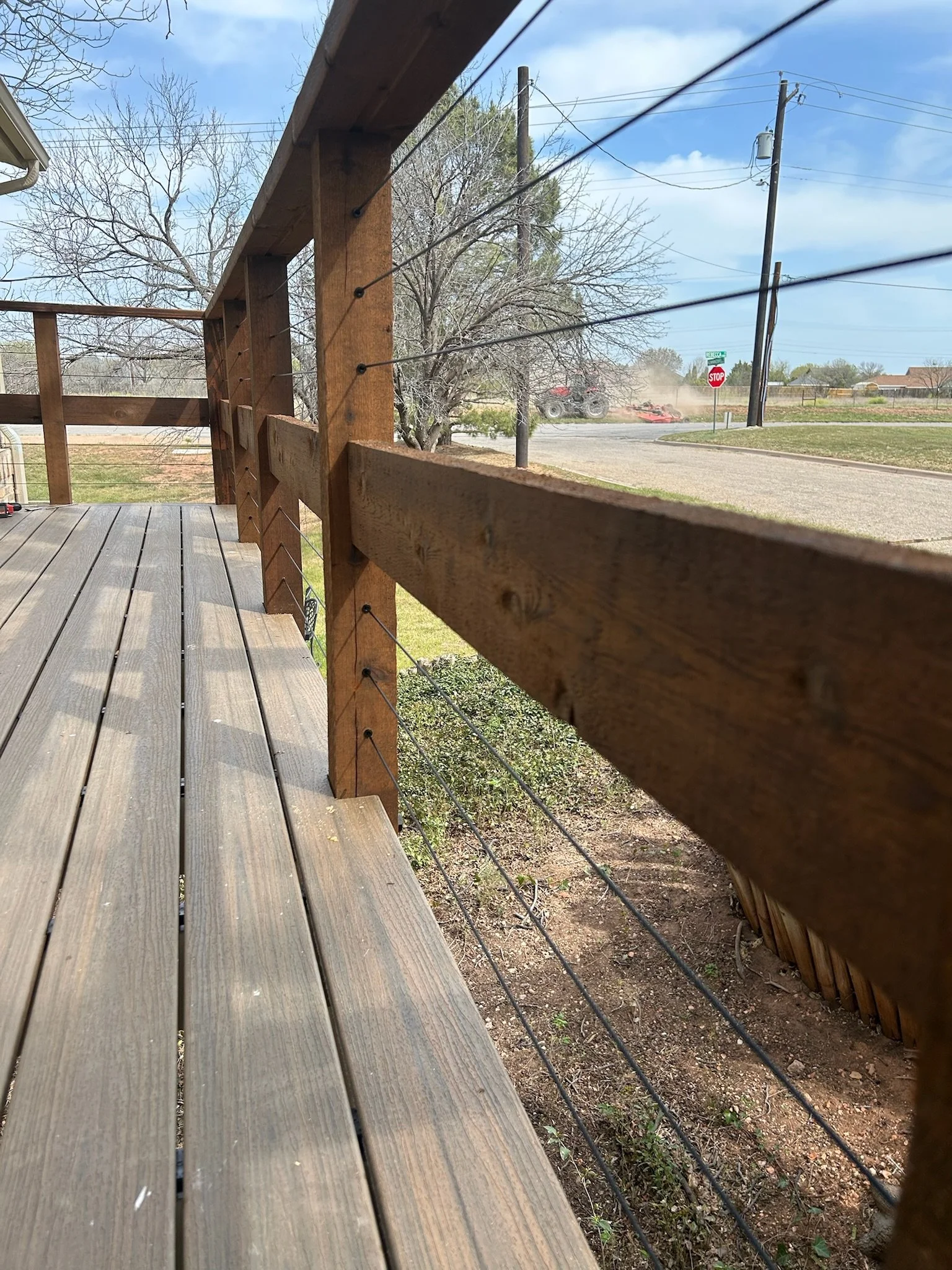 View of a wooden deck and railing, with a street scene in the background showing utility poles, trees, a stop sign, and a tractor with a dust cloud behind.