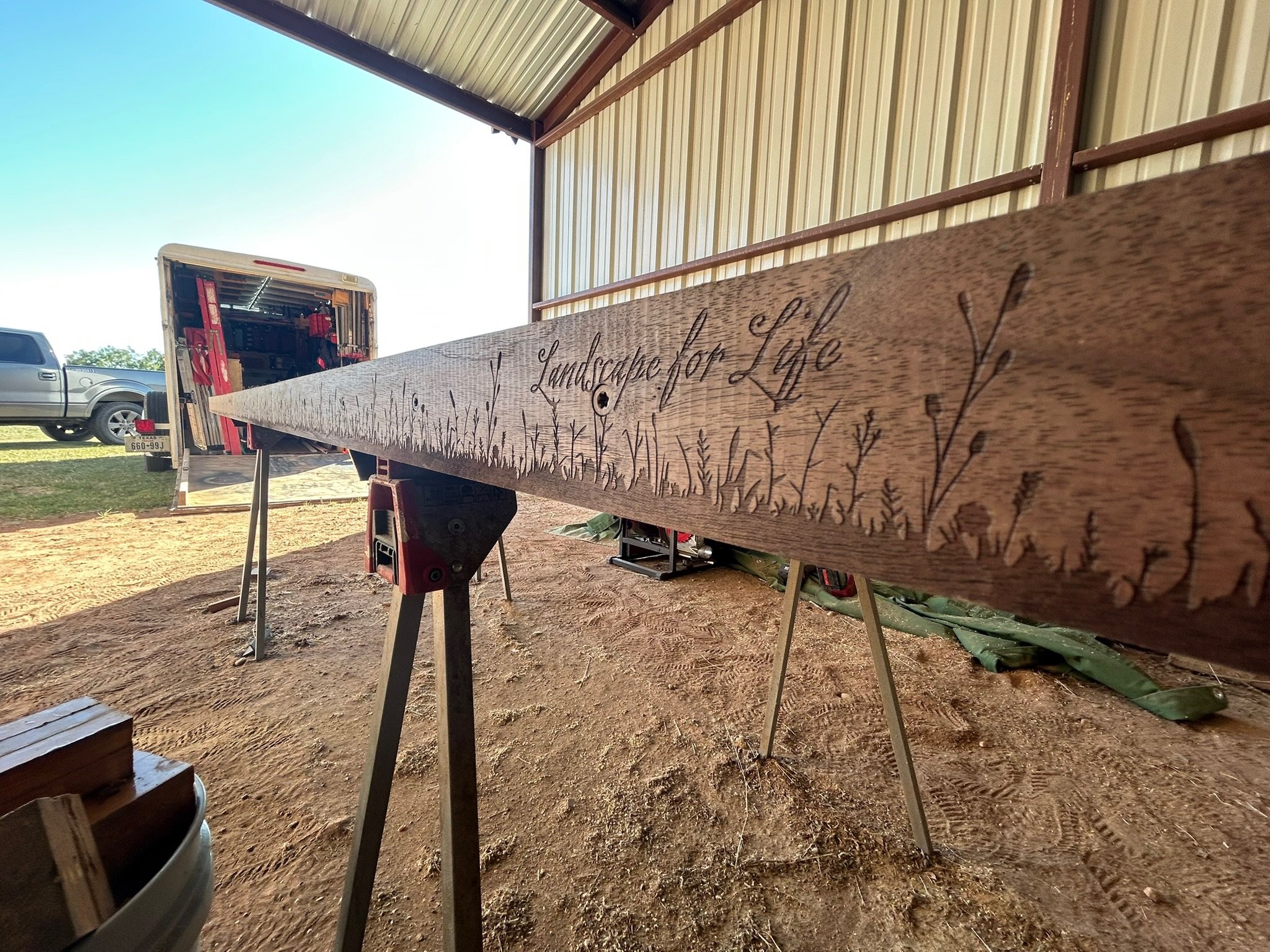 A wooden sign with the phrase 'Landscape for Life' engraved on it, positioned on saw horses inside a metal building or shed with a dirt floor and vehicles outside.