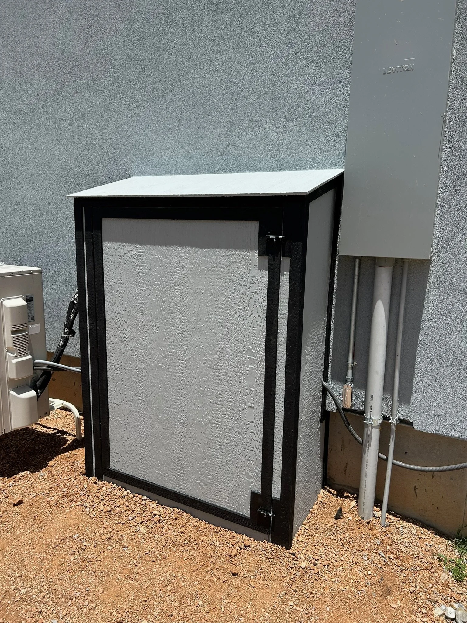 An electrical or utility box with a white reflective surface, black frame, and a latch on the side, installed on a cement wall. Piping and conduit are visible nearby, with the ground covered in loose gravel.