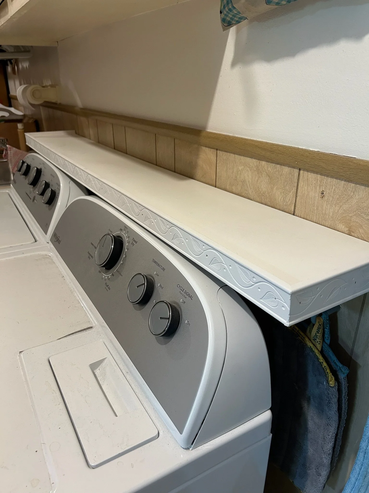 A white top-loading washing machine with buttons and dials, situated in a laundry area with a beige countertop and backsplash. There are some objects on the countertop, including a bottle of laundry detergent and a cloth.