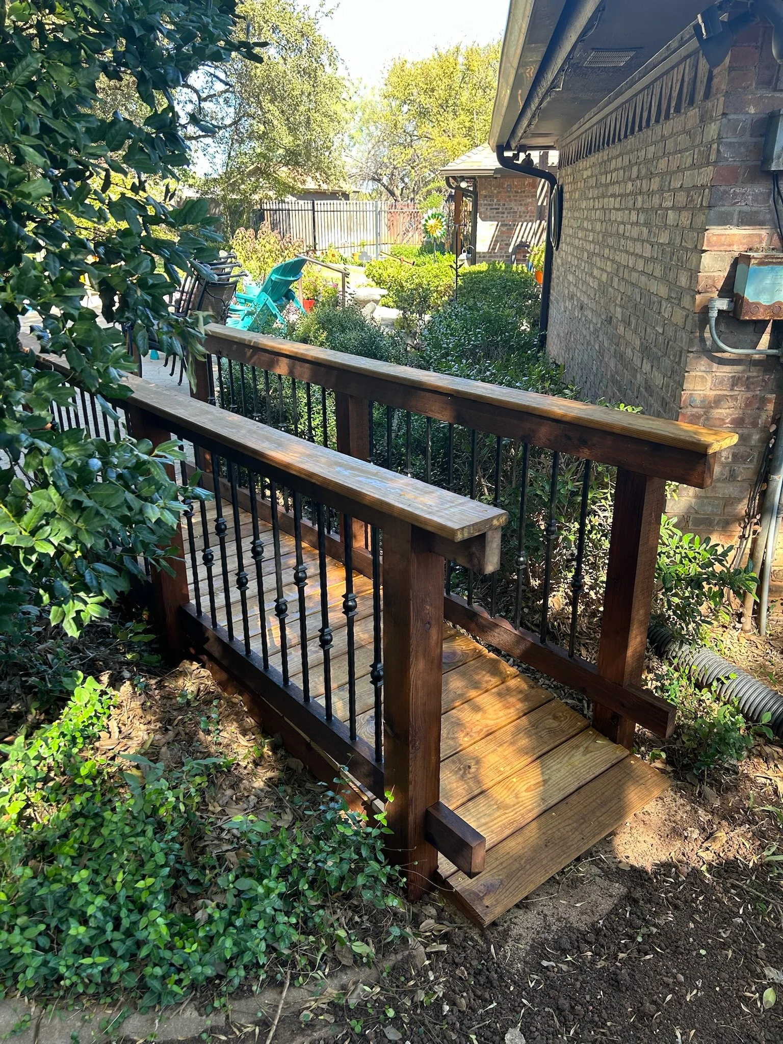 A newly built wooden deck with a railing, situated on the ground near a brick house, surrounded by bushes and trees, with outdoor chairs and a wind spinner in the background.