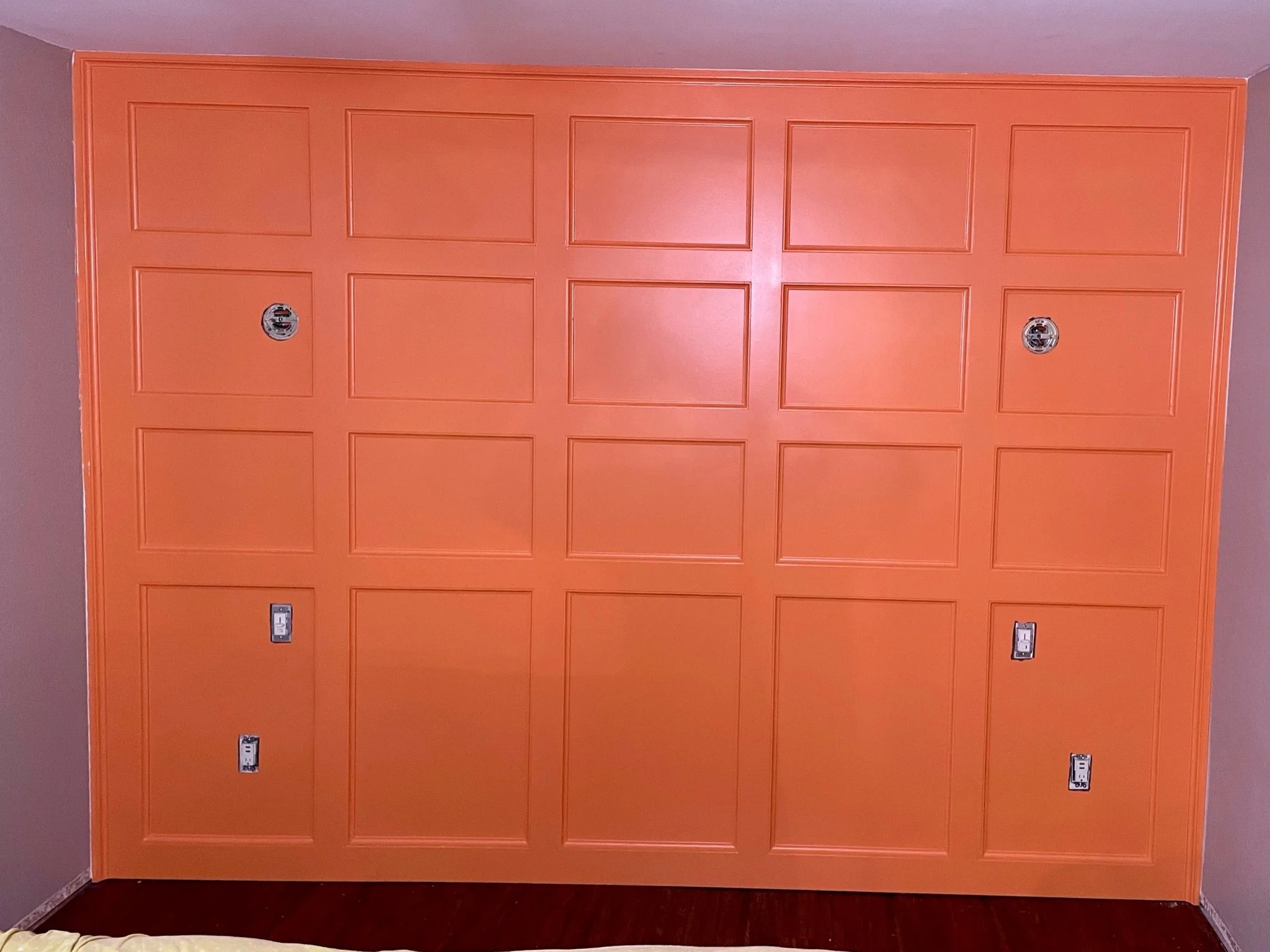 Orange wall with six square panel sections, two electrical outlets, and four uncovered electrical boxes.