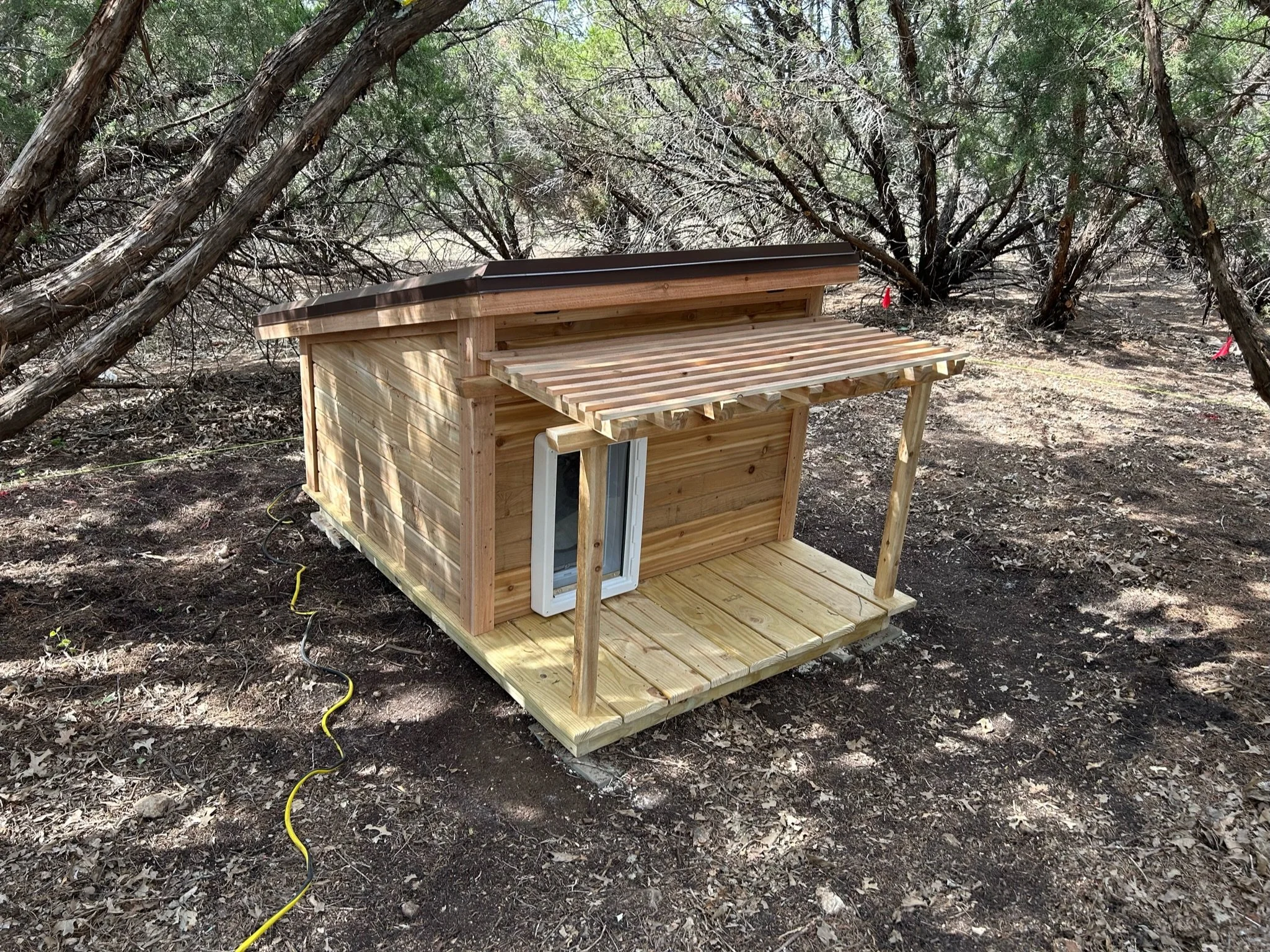 A small wooden doghouse with a sloped roof, situated in a wooded outdoor area with trees and dirt ground.