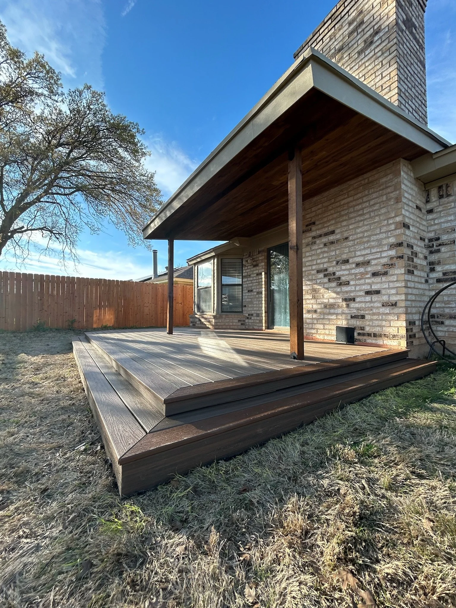 Backyard patio with wooden deck, covered by a roof supported by wooden beams, brick house exterior, bay window, surrounded by grass and a wooden fence, with a large tree nearby and a blue sky overhead.