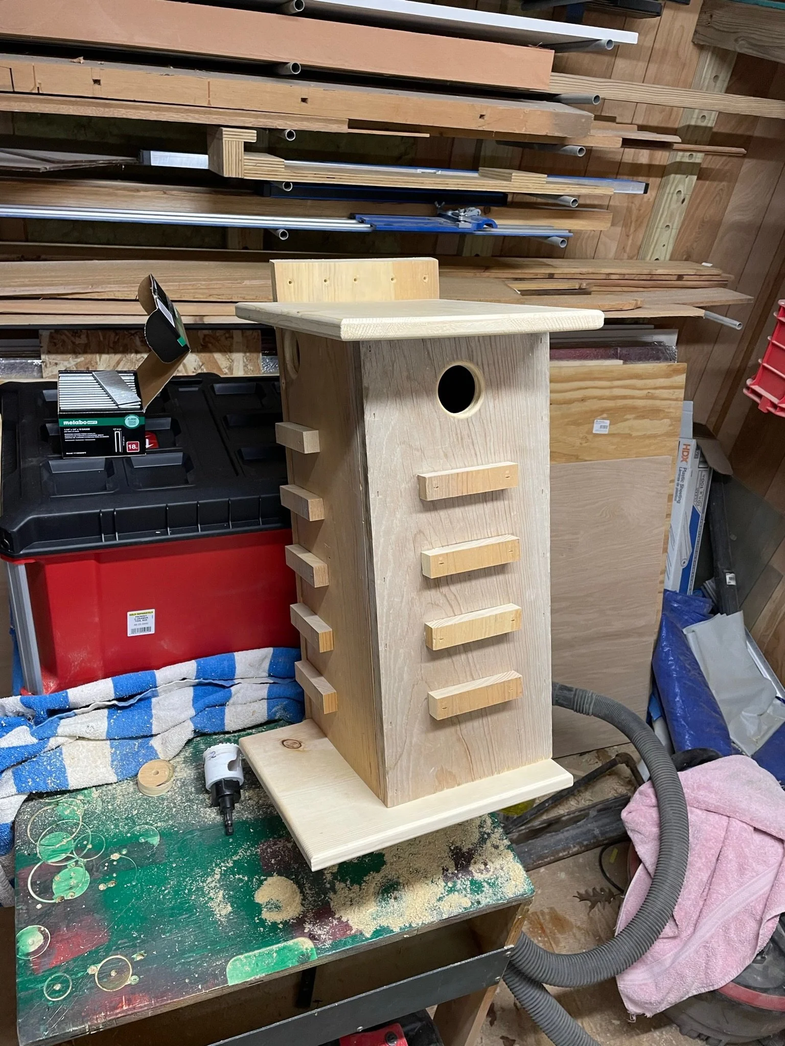 A workbench in a woodworking shop with a partially assembled wooden birdhouse, various tools, and materials scattered around, including a vacuum hose, a towel, and a power drill.
