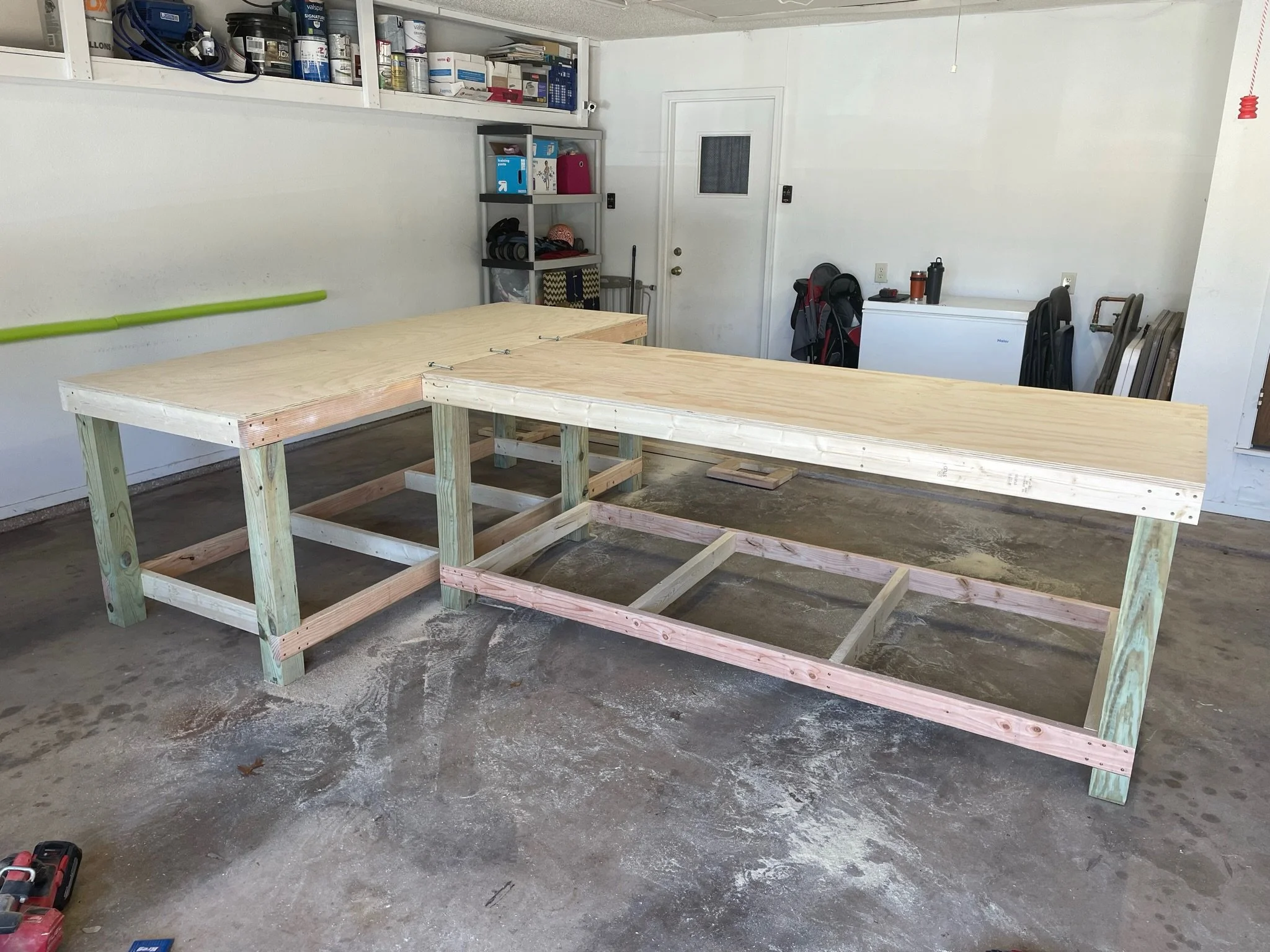 A garage with unfinished wood tables on a concrete floor, filled with tools, storage shelves, and a small white freezer.