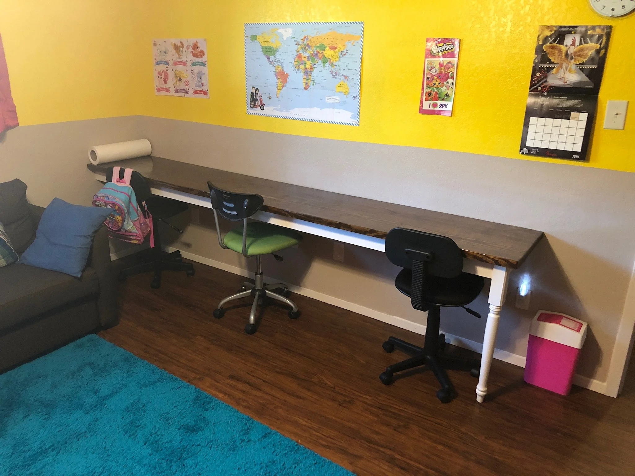 A classroom corner with a gray sectional sofa and various pillows, a long wooden desk with three chairs, and a yellow wall decorated with colorful posters including a world map, a calendar, and educational images, as well as a clock and a pink trash 