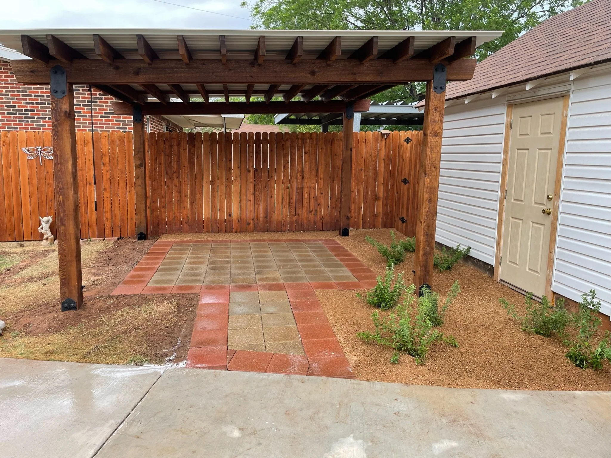 A backyard with a wooden fence, a covered patio area with tile flooring, small plants, and decorative butterflies on the fence. There's a white house with beige door and shingles on the roof.