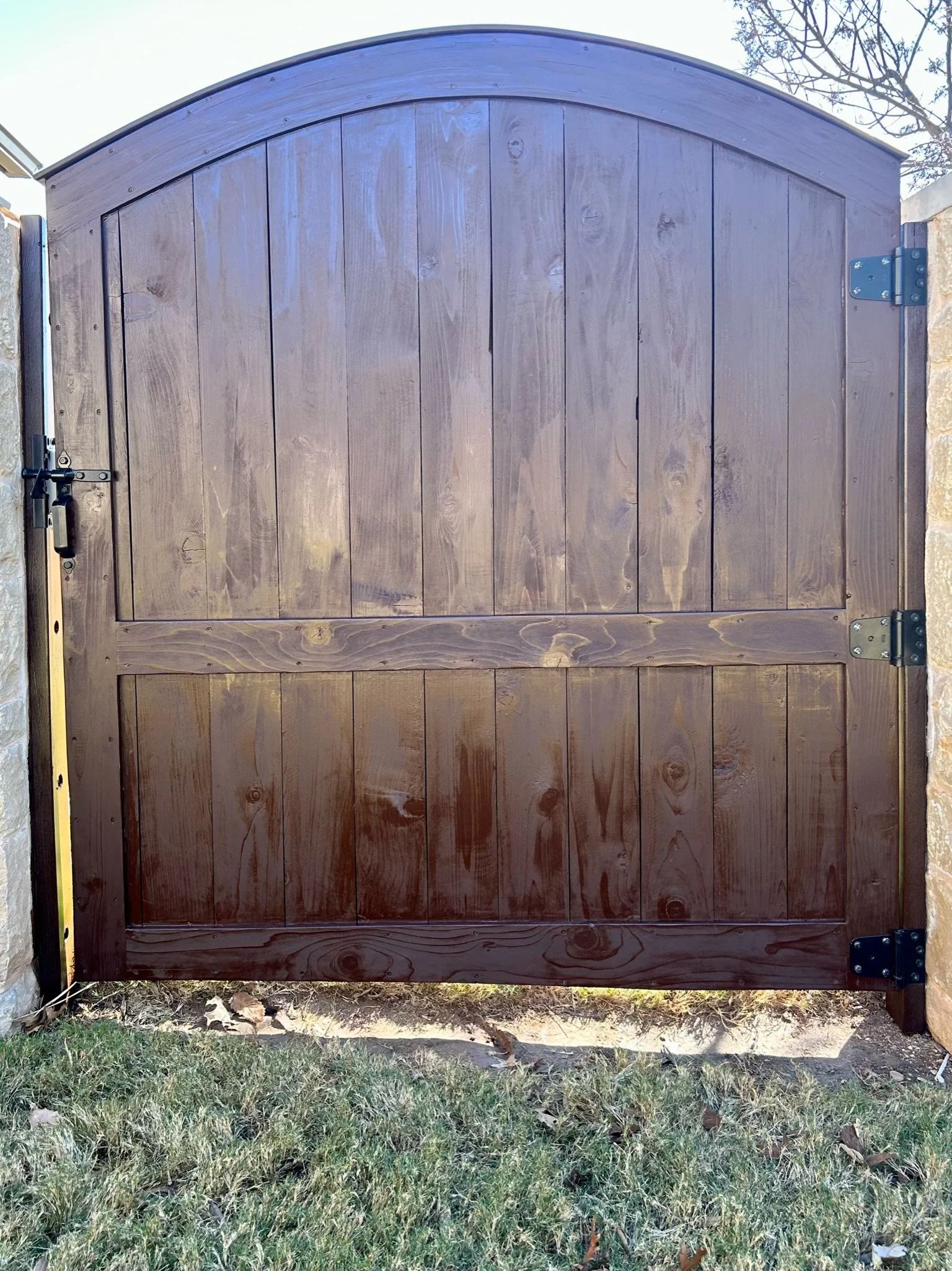 A wooden gate with metal hinges and a latch, standing on grass with some fallen leaves, part of a fence, with a tree and sky in the background.