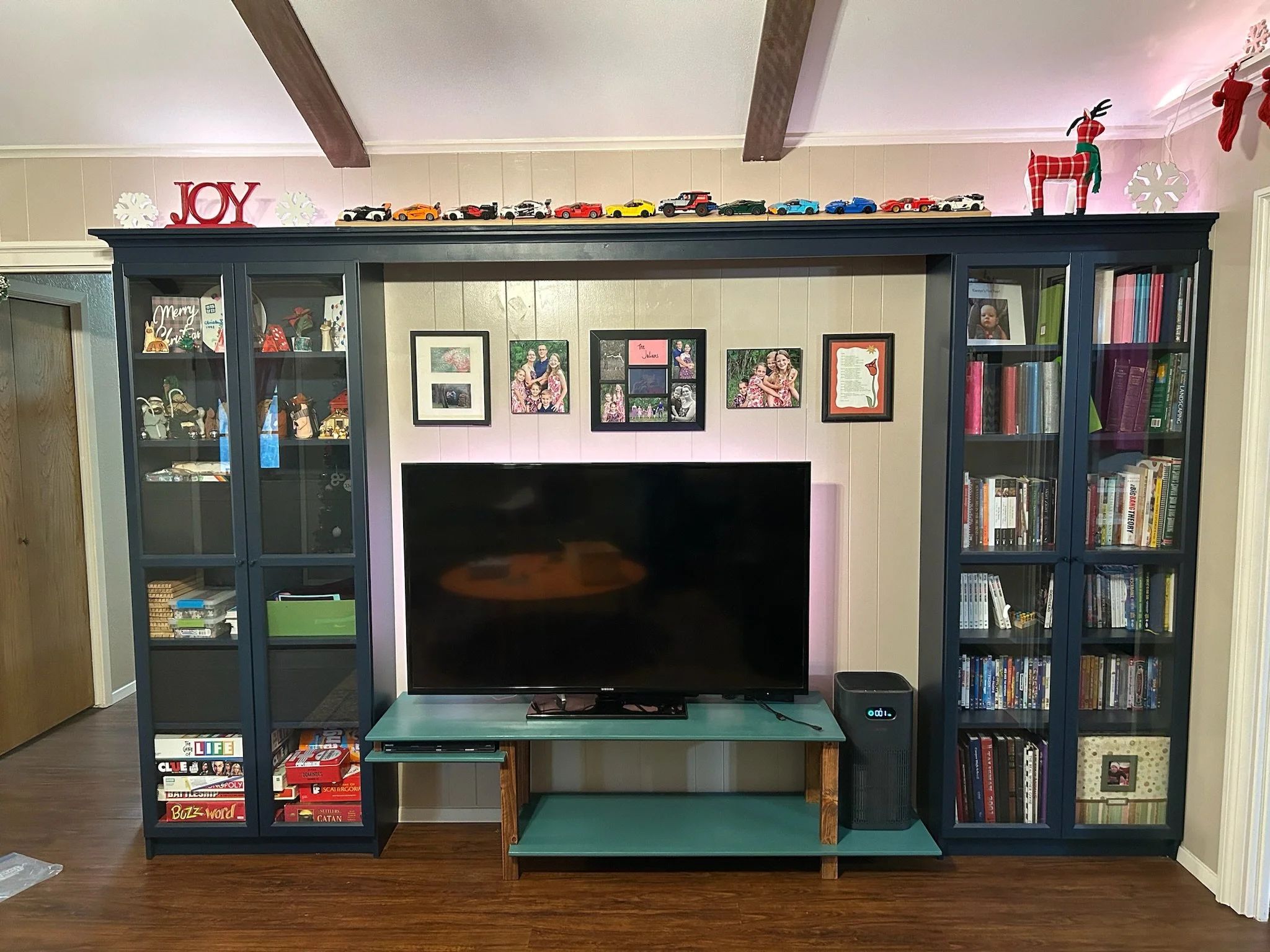 A dark blue entertainment center with a television on a teal stand in front of a beige wall with framed family photos. Decor includes a red 'JOY' sign and Christmas decorations, including a red and white plaid reindeer, snowflakes, and a red stocking