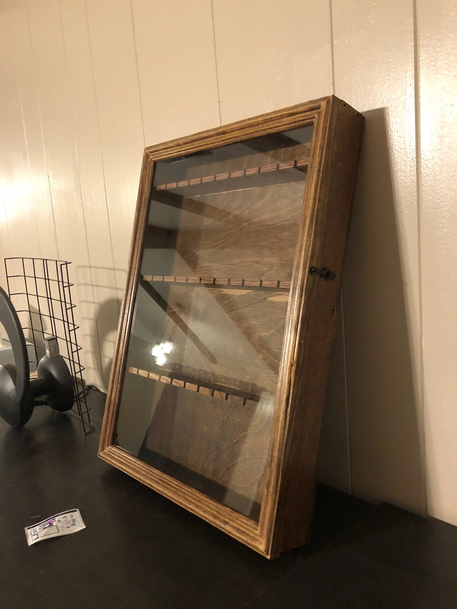 A wooden display cabinet with glass door, empty inside, leaning against a beige wall in a room with black floor. To the left, a black wire rack and a small bundle of cables or wires are visible on a black surface.