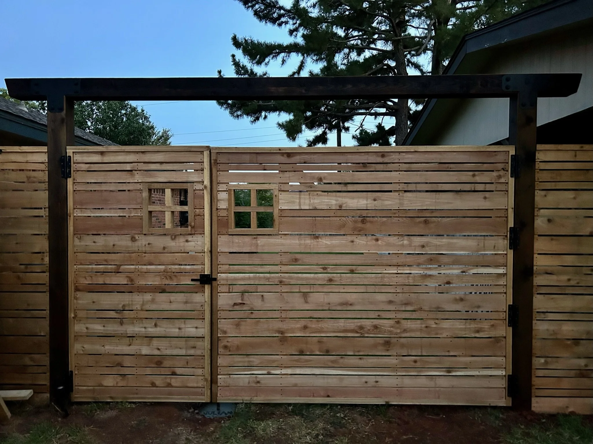 A wooden fence with horizontal slats and a small square window in the middle, supported by dark brown posts, with a house roof and trees in the background under a clear sky.