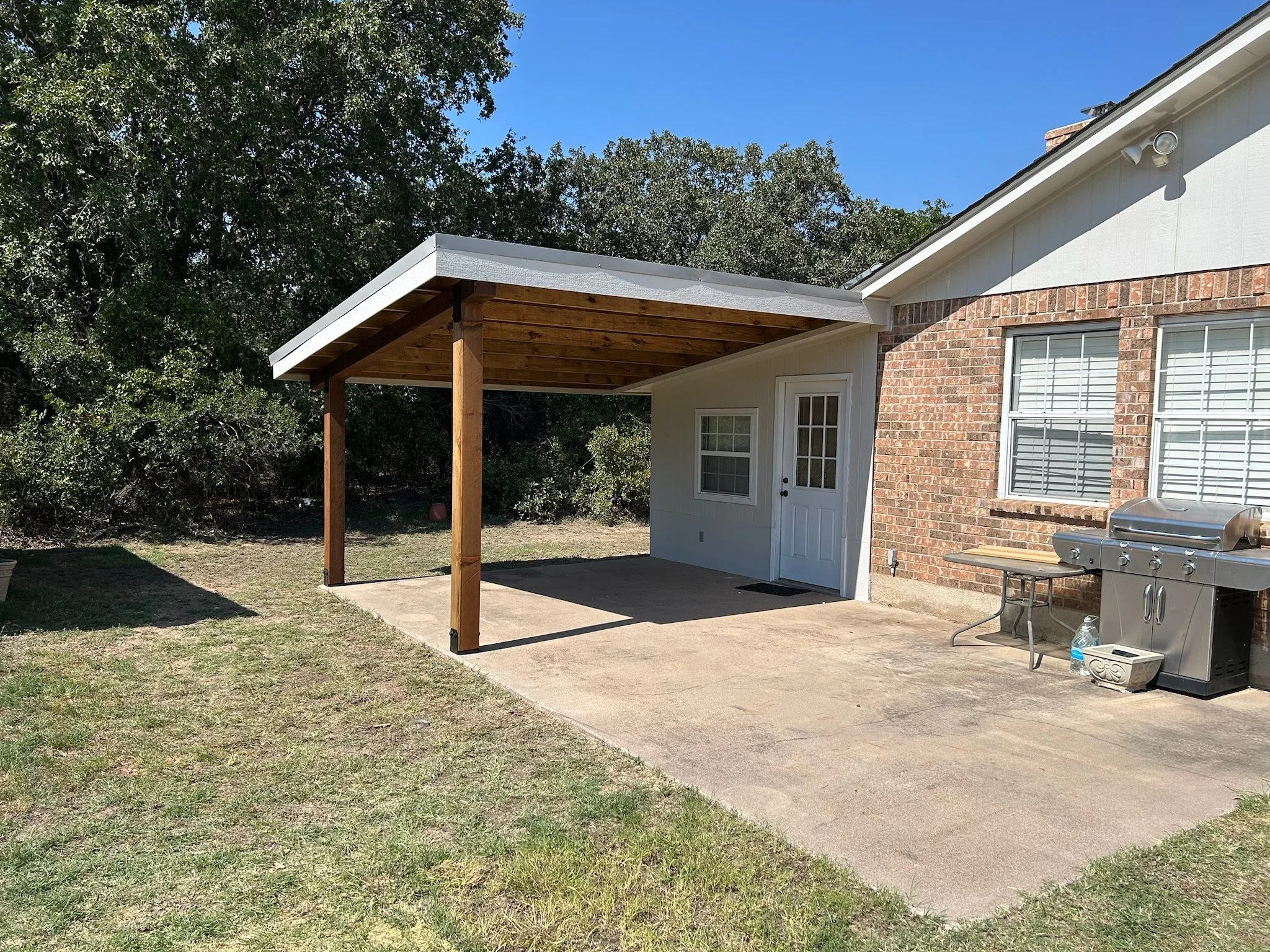 A backyard patio area with a concrete slab, a wooden roof extension supported by wooden posts, a brick house with windows and a door, a stainless steel grill, and a gravel area with some grass and trees in the background.