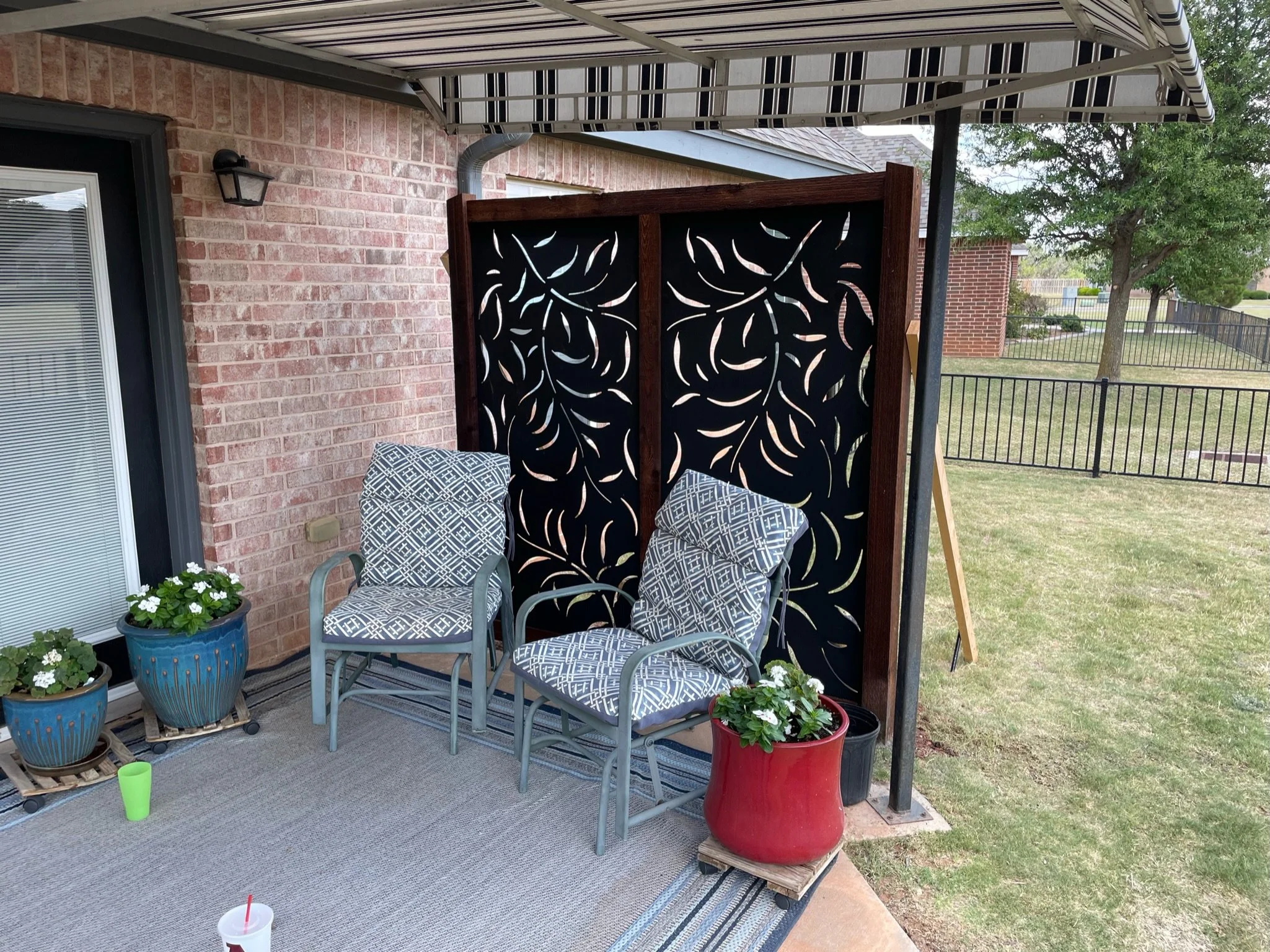 A patio area with a black decorative divider, two chairs with patterned cushions, multiple potted plants, and an outdoor rug. There's a brick wall in the background and a fence in the distance.