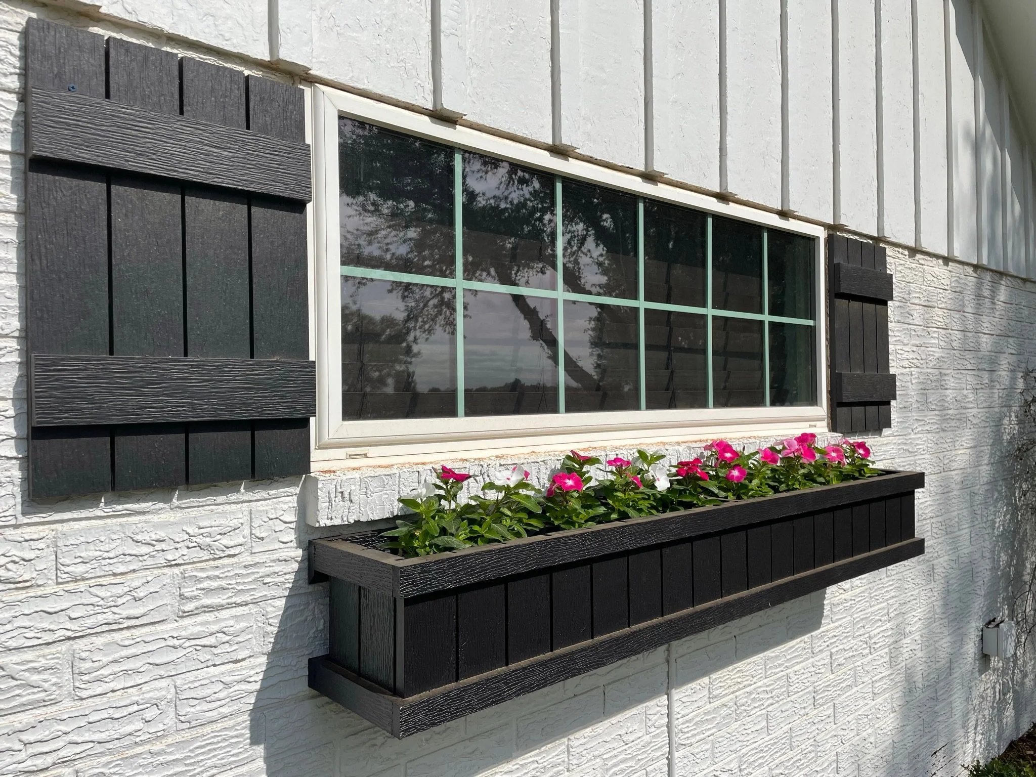 A white house exterior with three black window shutters, a flower box with pink flowers beneath a large window, and reflection of trees in the window glass.