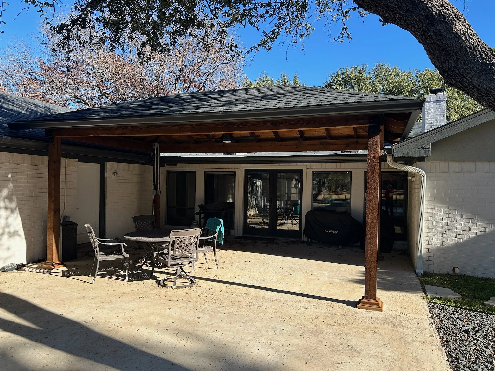 Backyard patio area with a wooden pergola, patio furniture including chairs and a table with a blue towel, surrounded by trees and blue sky.