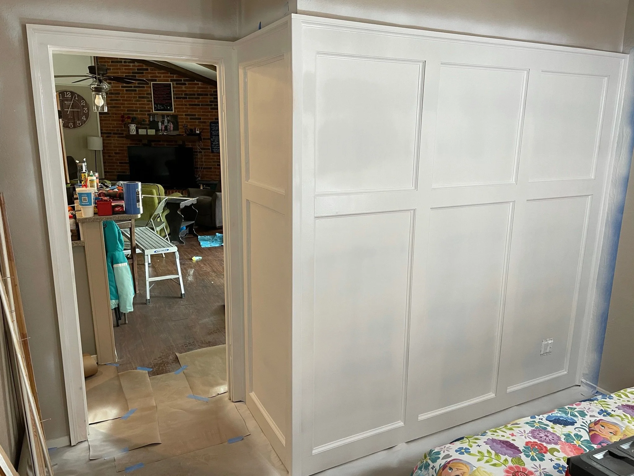 A partially assembled white built-in closet structure in a bedroom, with paper and painter's tape on the floor, and a colorful bedspread with a floral pattern visible in the foreground.