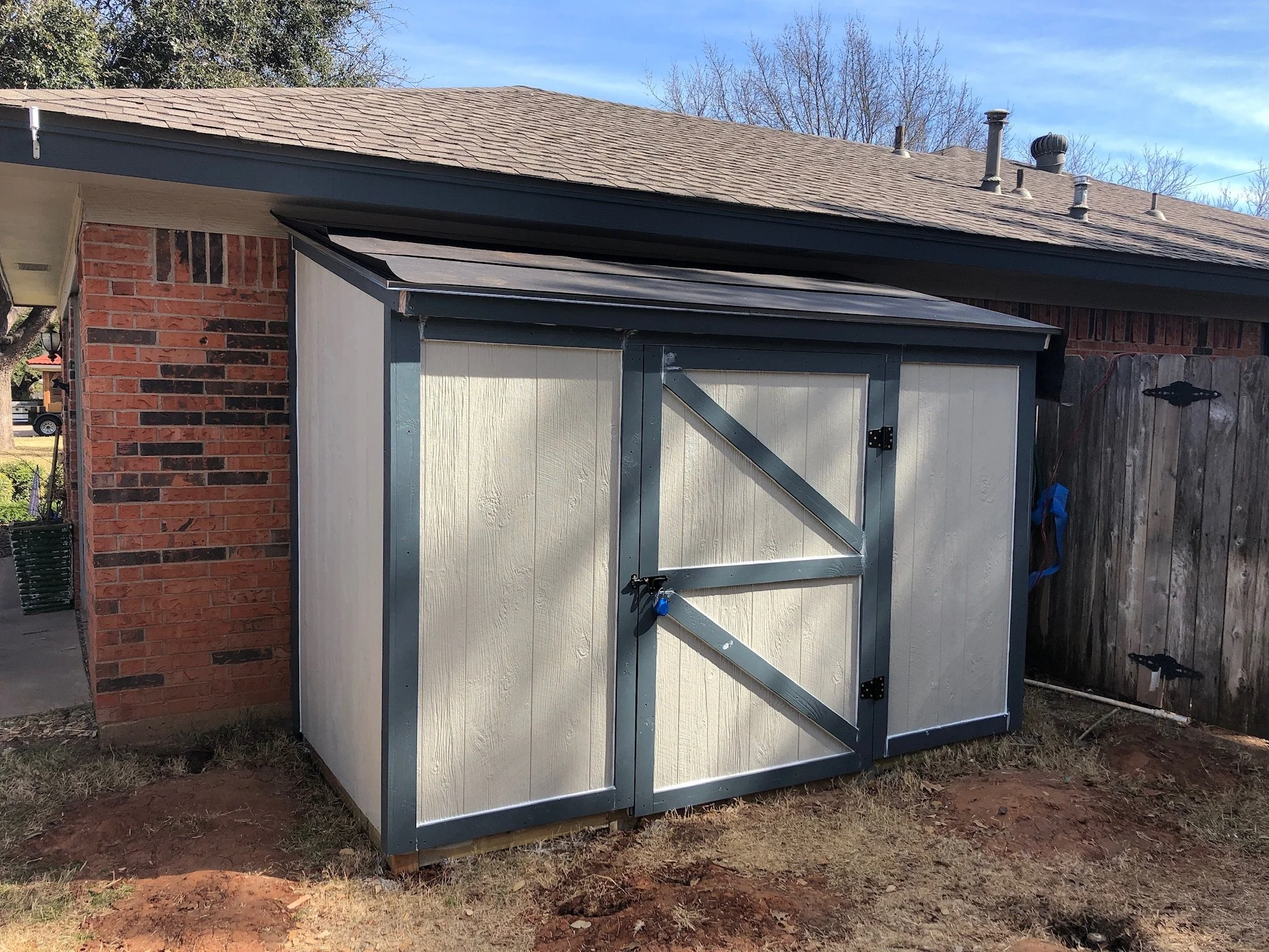 A small, gray shed with double doors, situated next to a brick house. The shed is on a patch of dirt and is enclosed by a wooden fence. The house has a brown shingled roof, with a brick exterior wall partially visible. The sky is clear with a few clo