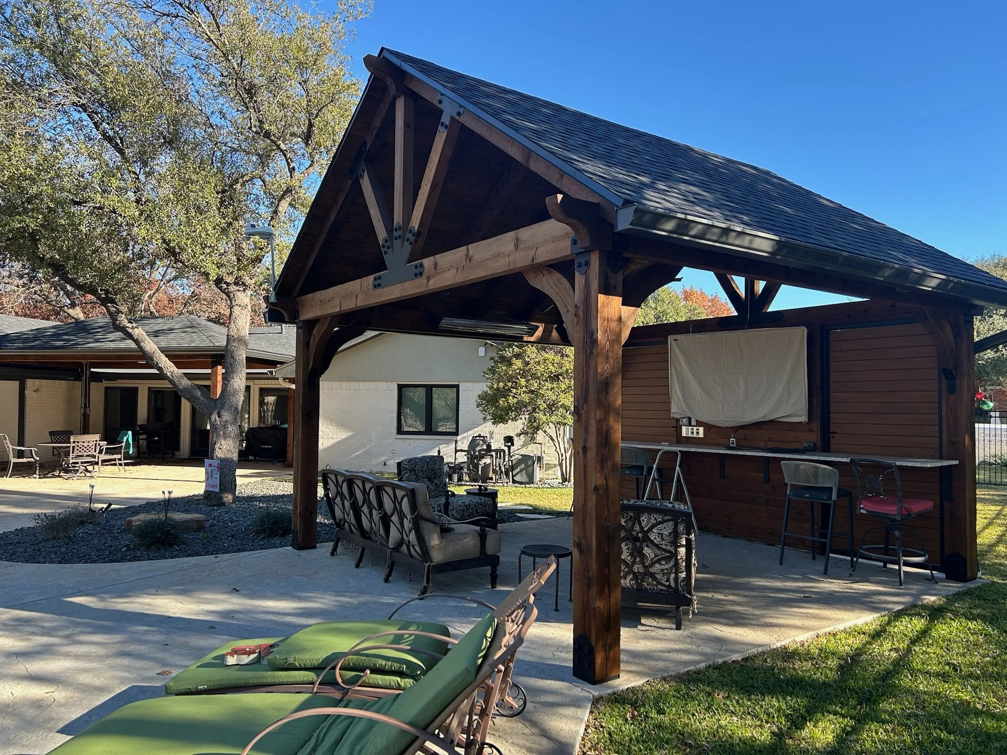 A backyard patio with outdoor furniture including chairs and cushions, a wooden structure with a sloped roof, trees with autumn leaves, and a clear blue sky.