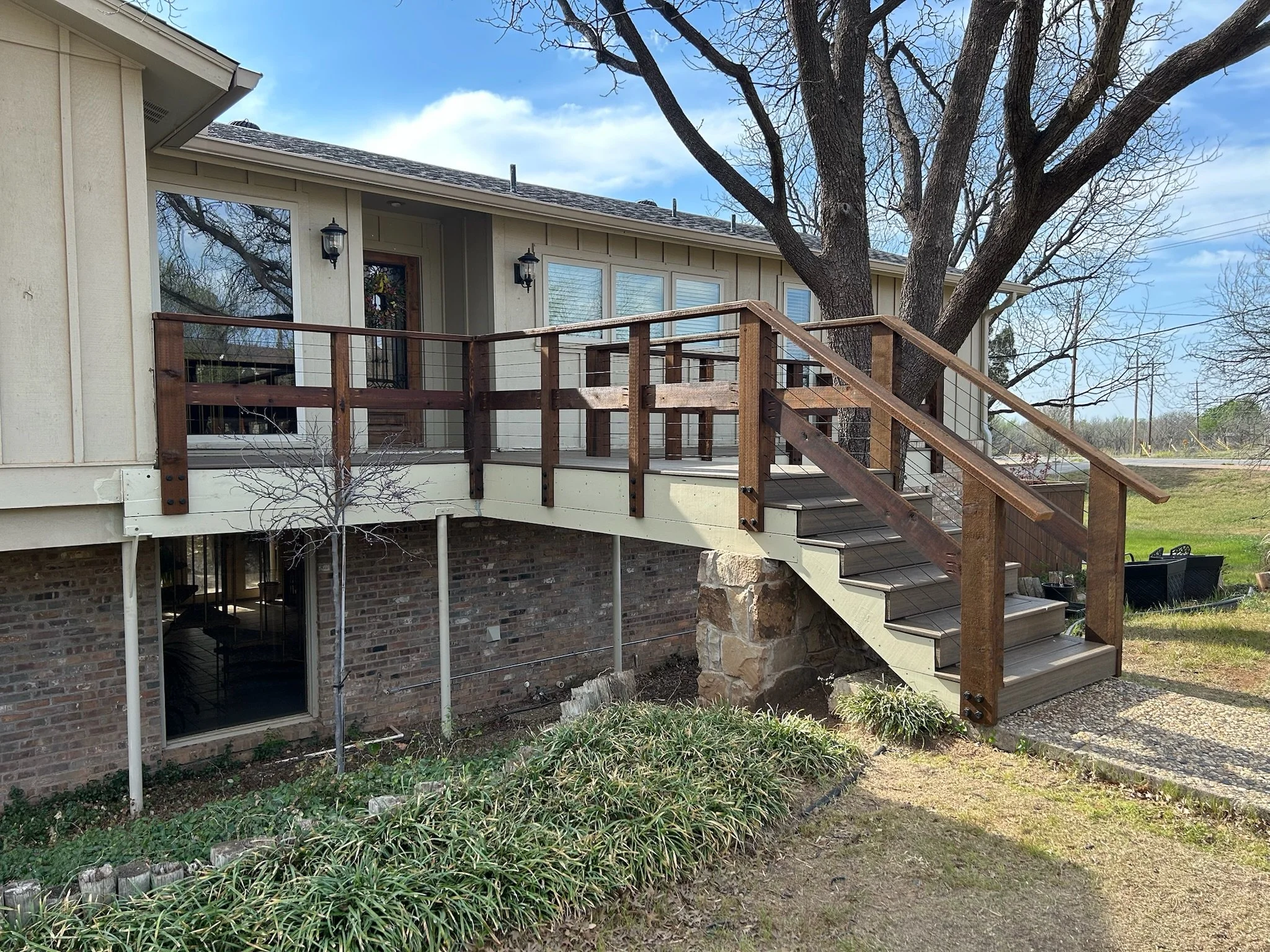 Photo of a residential house's backyard deck with stairs, surrounded by trees and greenery, under a partly cloudy sky.