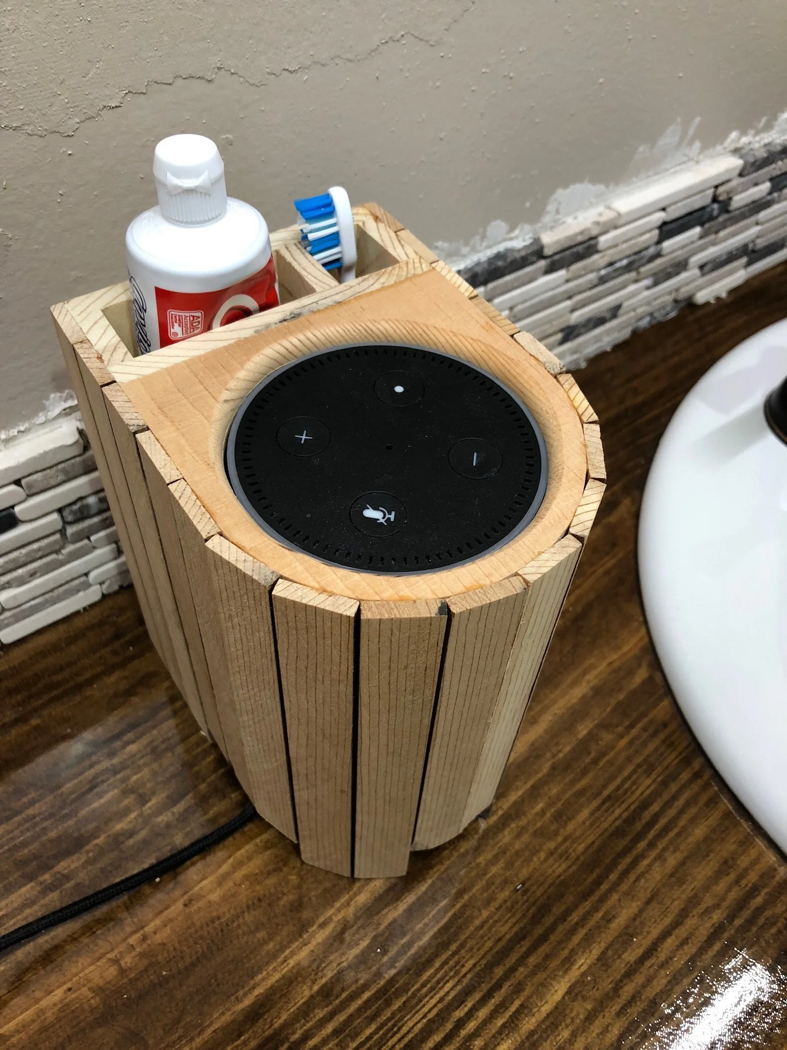A wooden stand with a black smart speaker on top, a bottle of glue, and a toothbrush in a holder, near a bathtub and a textured wall.