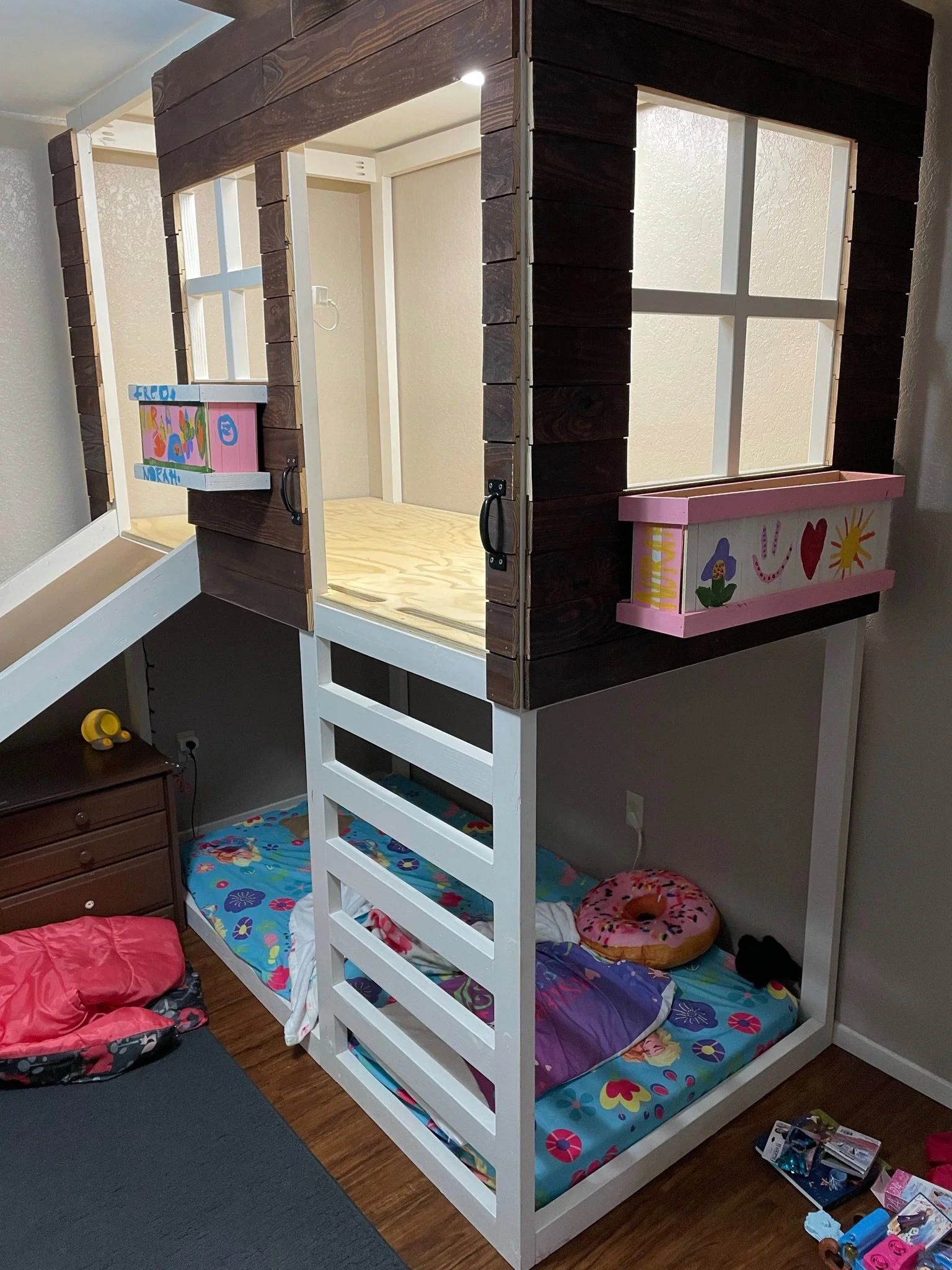 A children's bedroom with a white bunk bed, a queen-sized bed with colorful floral and purple bedding on the bottom, decorated with a donut pillow, and a small wooden nightstand with drawers. There are toys and books scattered on the floor and a smal