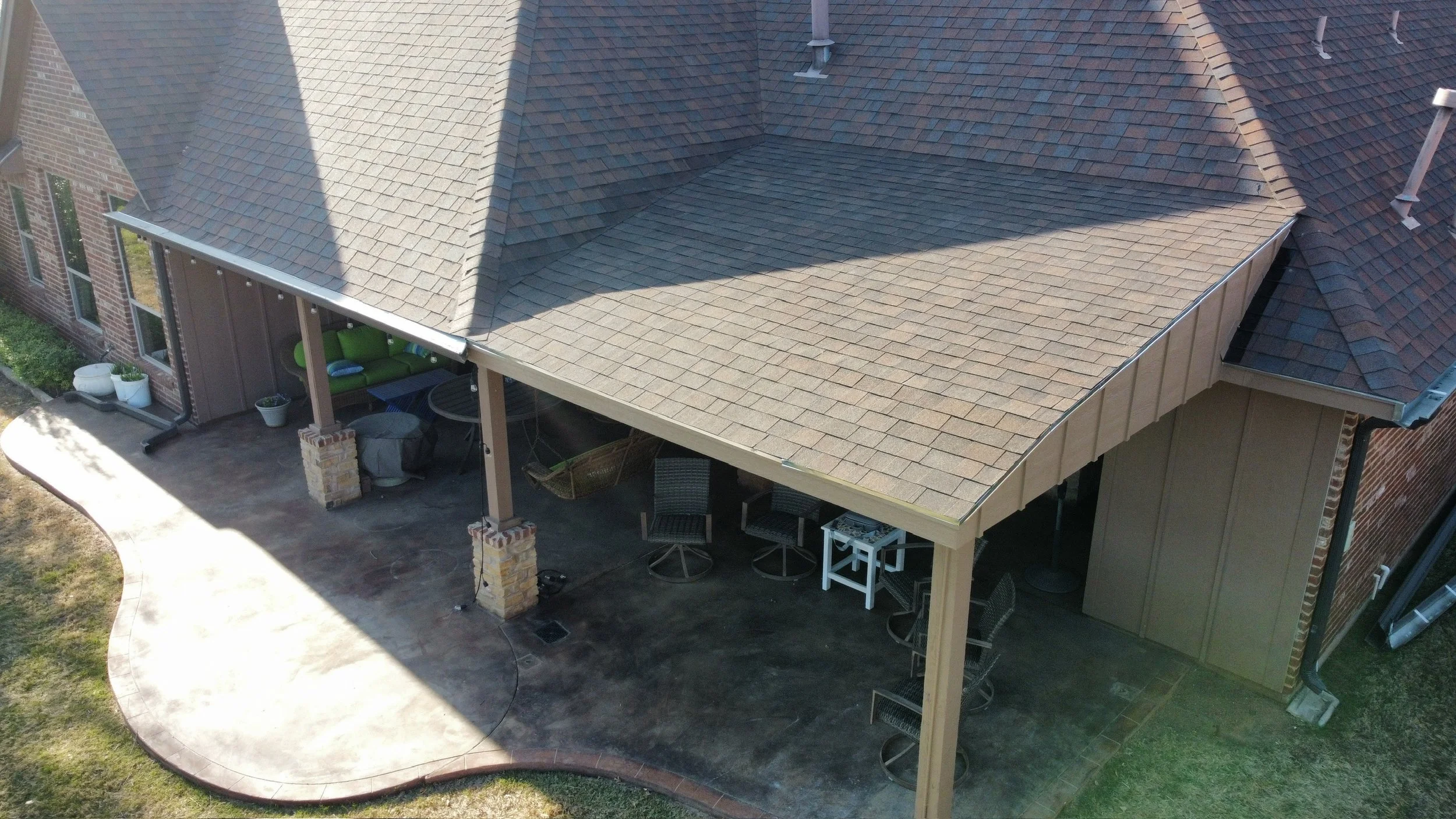 Aerial view of a house porch with a large, tan-colored, shingled gable roof, brick walls, and an outdoor seating area underneath a covered section with chairs, tables, and potted plants.