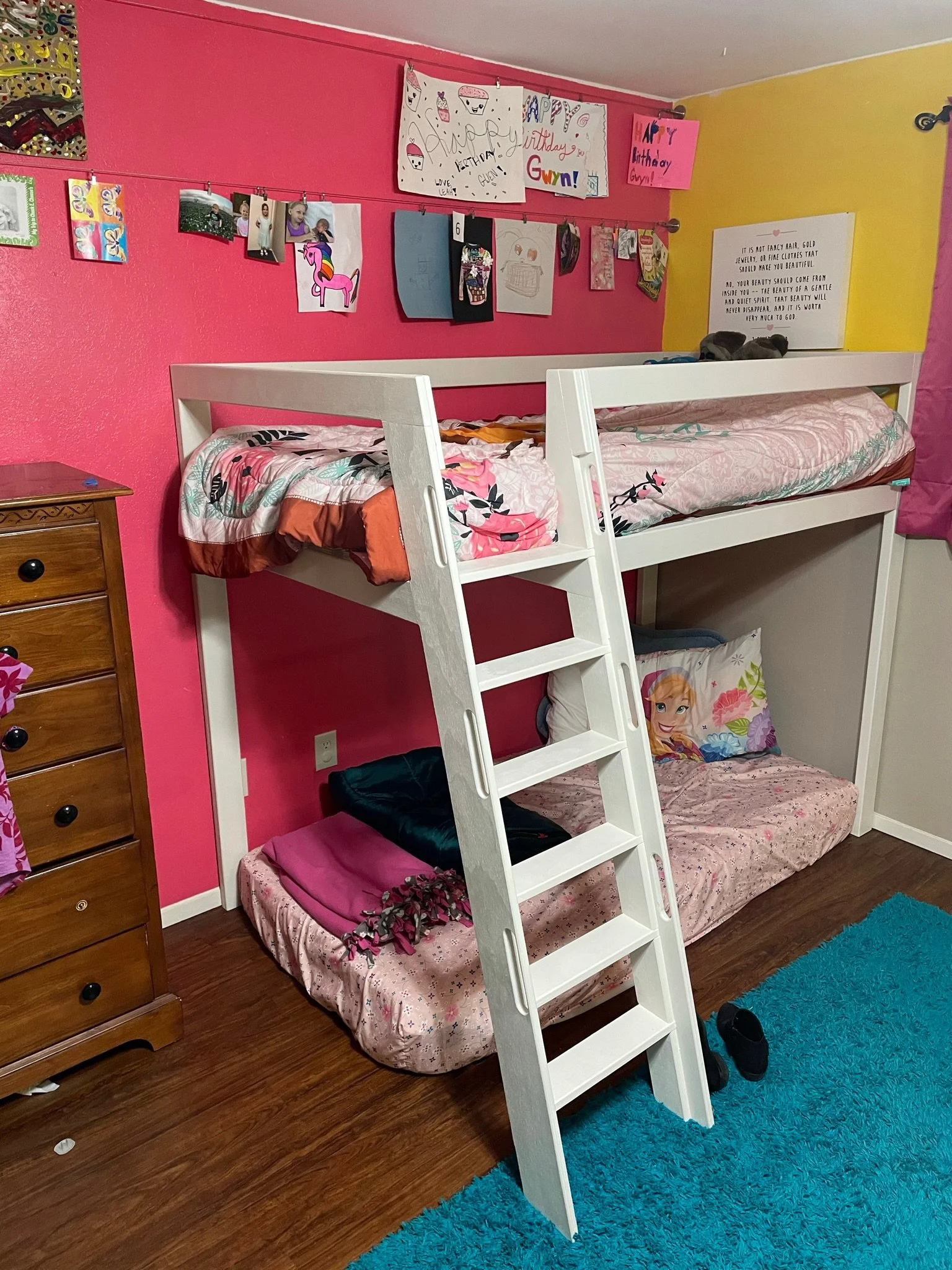 A cozy bedroom with pink and yellow walls featuring a white bunk bed with a ladder. The top bunk has floral bedding, and the bottom bunk has a Princess Anna pillowcase from Frozen. A small pink mattress with bedding and a bed of artificial flowers is