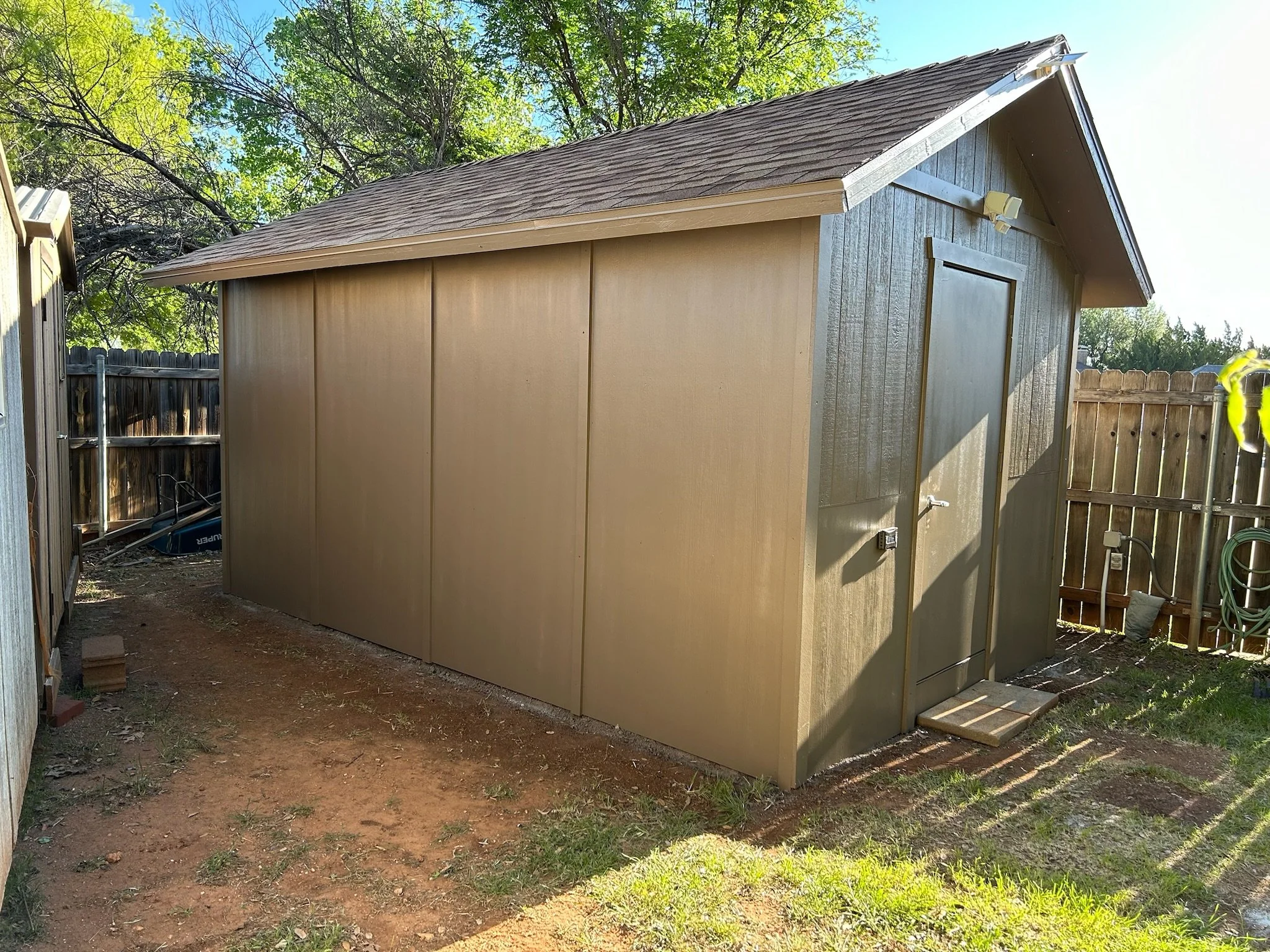 A beige shed with a gable roof in a backyard surrounded by a wooden fence and trees with green leaves. There is a small step made of wood in front of the shed door.