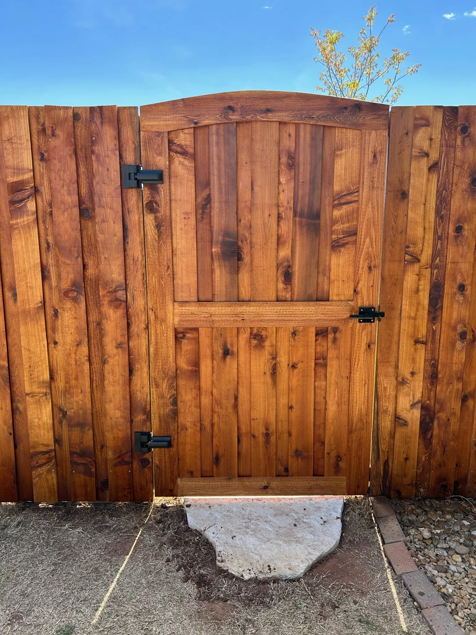 A wooden gate with hinges, installed against a backdrop of clear blue sky and a small tree. The gate is supported by a concrete foundation with a large rock at the base and some surrounding soil and gravel.
