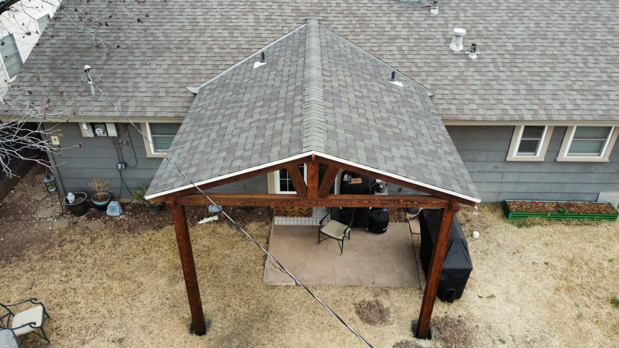 Aerial view of a house with a gray shingle roof, an attached porch with wooden support beams, a patio with outdoor furniture, potted plants, and a grill, and surrounding yard with grass and gravel.
