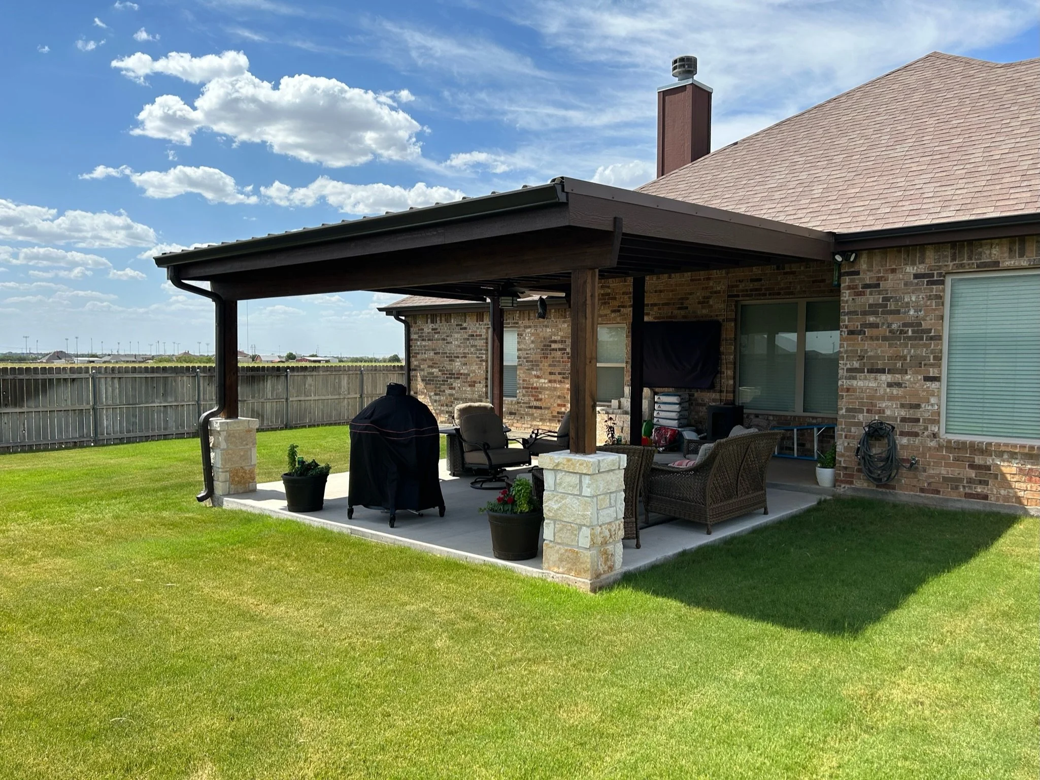 Backyard patio area with outdoor furniture under a covered patio, brick house with a chimney, and a wooden fence in the background, under a blue sky with scattered clouds.