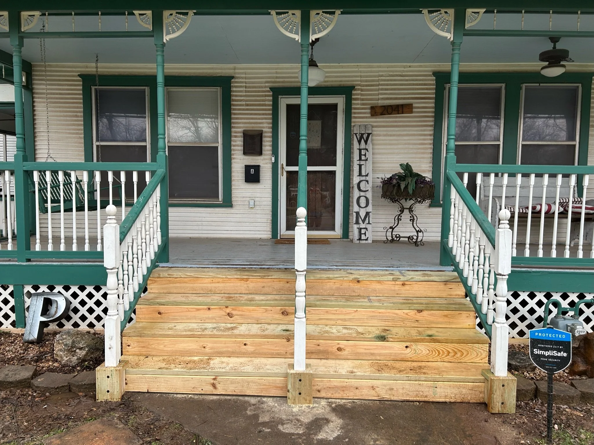 Front porch of a house with a wooden staircase, white railings with green accents, and a welcome sign. The house has beige siding, green window trims, and a porch ceiling. A potted plant is to the right of the door, with two window air conditioning u