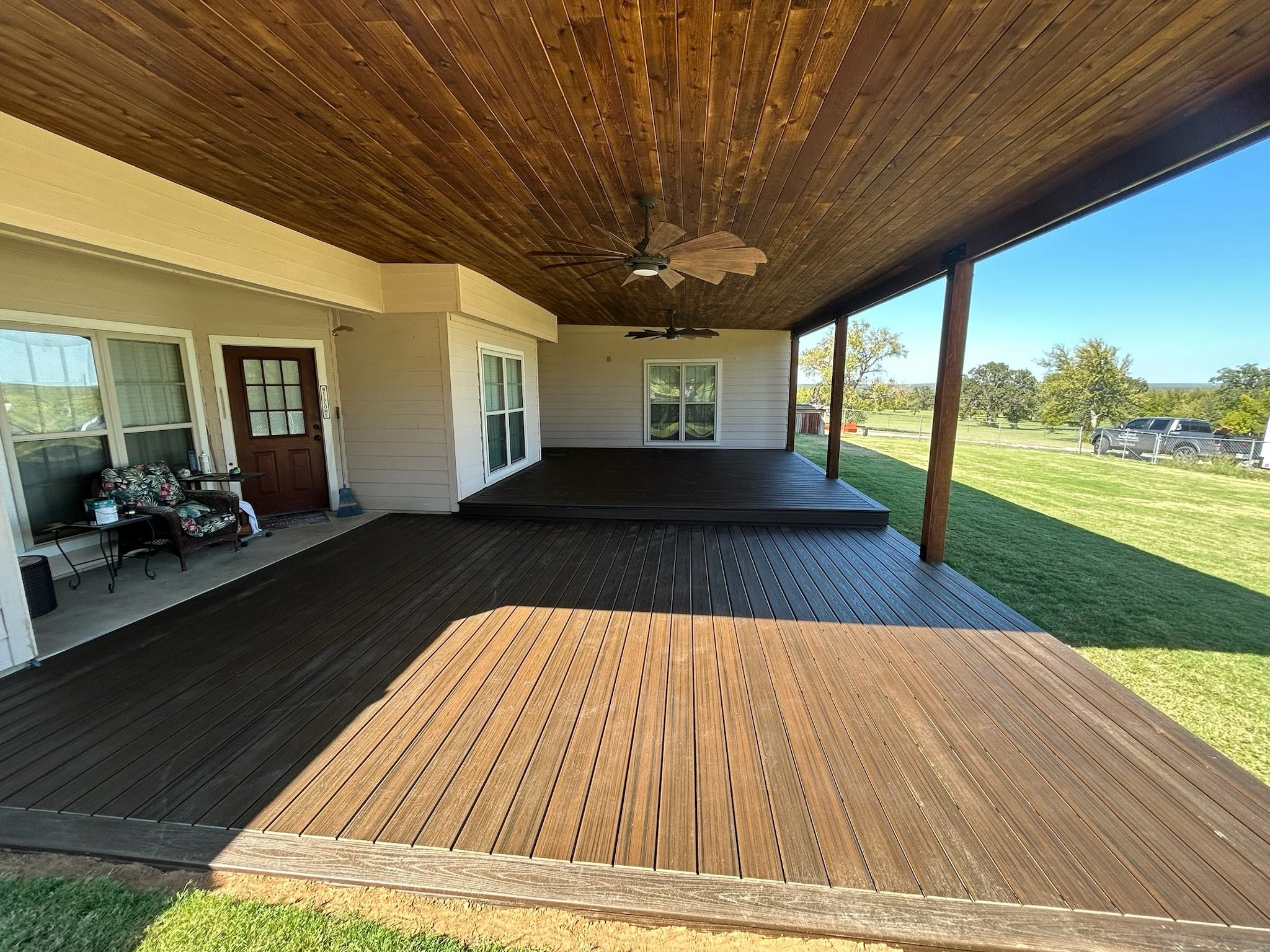 Covered porch with wooden ceiling, two ceiling fans, beige siding, two windows, and a door, overlooking a green yard with trees and a car.