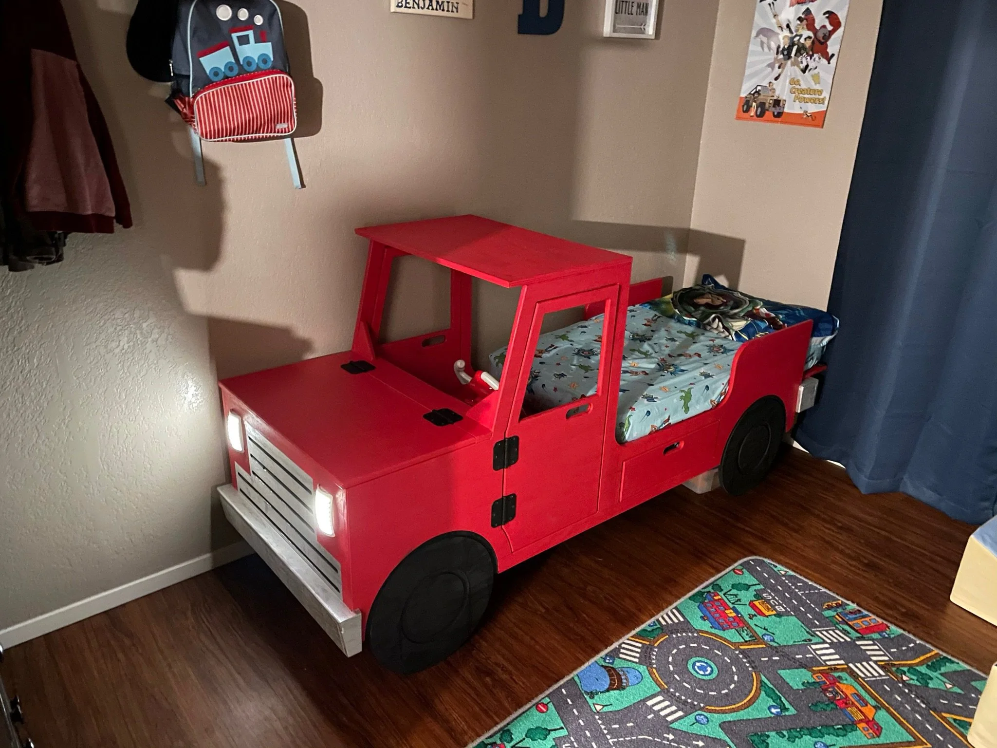 A children's bedroom corner featuring a red truck bed with a colorful dinosaur print mattress, a blue and black rug with a city street pattern, and a hanging backpack with a train design on the wall.