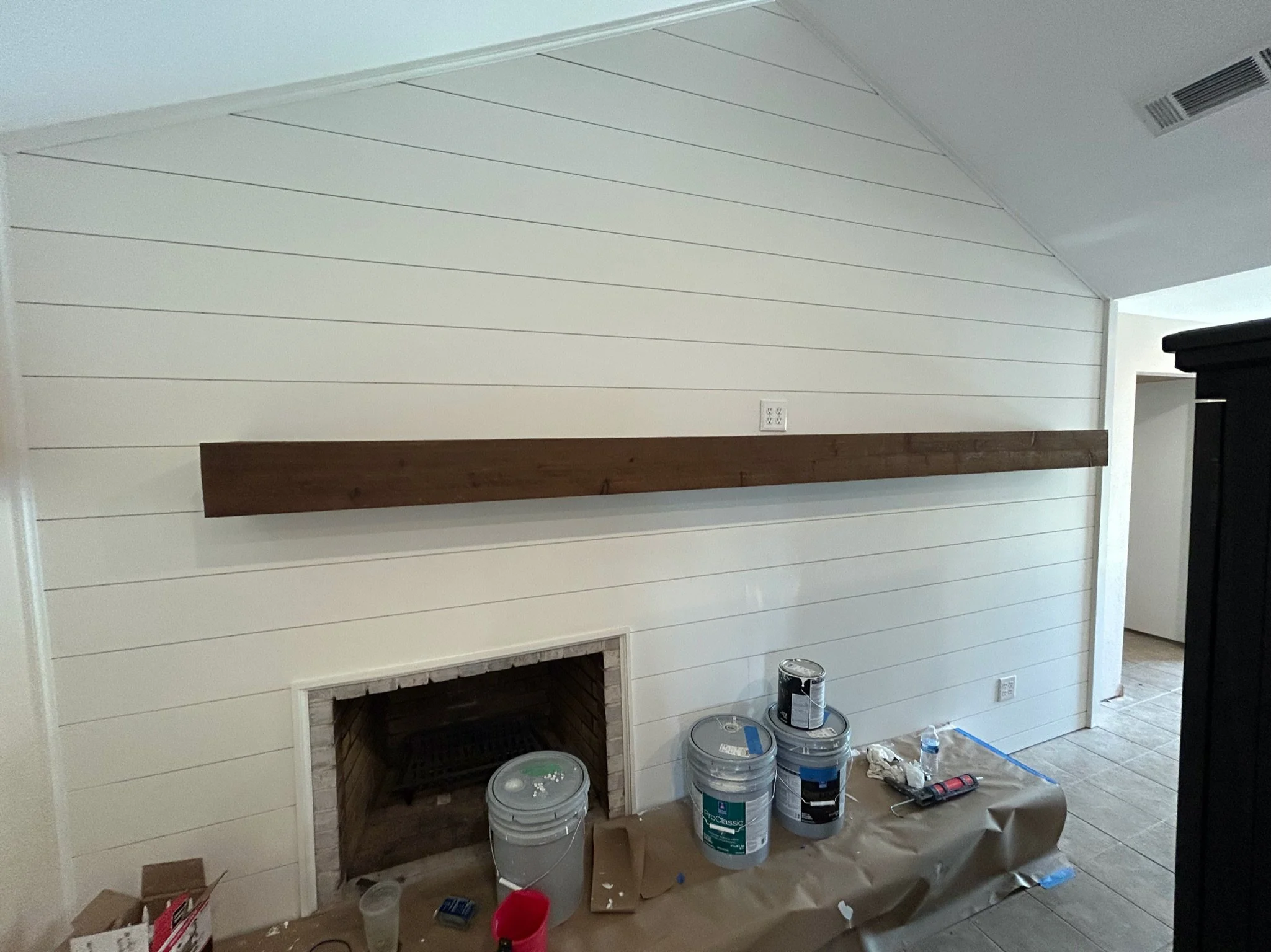 Interior of a living room under renovation with a new white shiplap accent wall, a fireplace opening with brick inside, and painting supplies on the floor including several paint cans, brushes, and painter's tape.