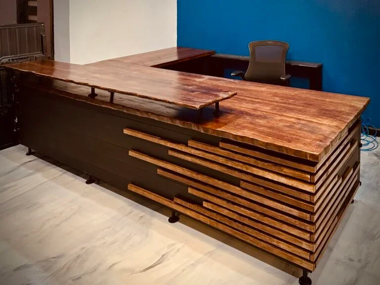 A large wooden desk with a unique, layered design featuring uneven, horizontal wooden slats, set in an office with a chair and a blue accent wall.