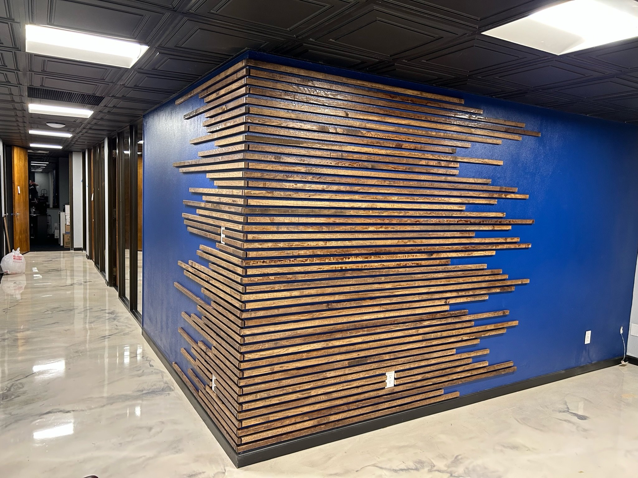 Interior view of a modern building with a blue wall feature that has horizontal wooden slats arranged in a wave pattern. The ceiling has black panels with recessed lighting, and the floor is shiny marble.