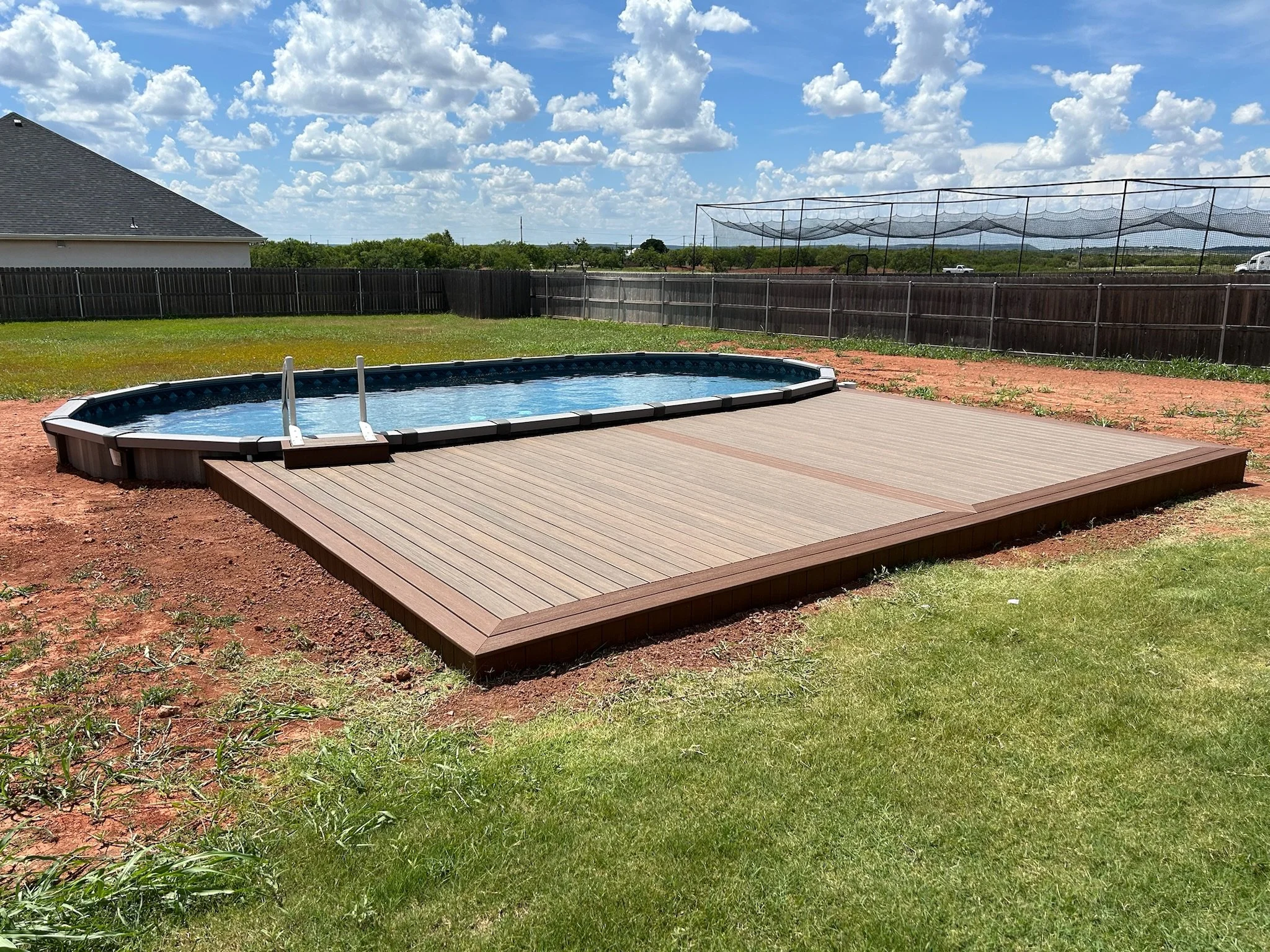 Backyard with a small above-ground swimming pool on a wooden deck, surrounded by green grass and a wooden fence, under a partly cloudy sky.