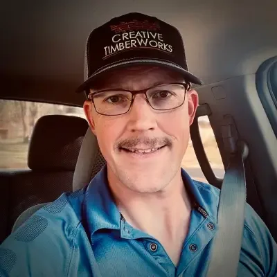 Man wearing glasses and a black cap with 'Creative TimberWorks' logo, smiling inside a vehicle.