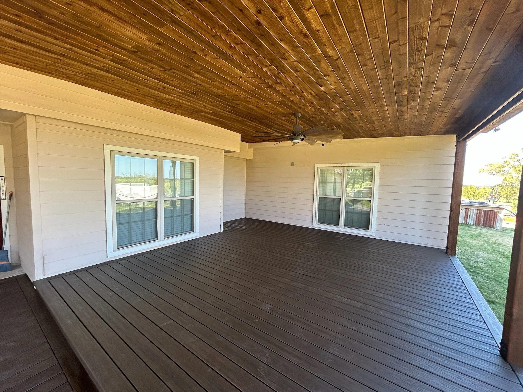 Covered outdoor patio with wooden ceiling, ceiling fan, and sliding glass doors to the interior of a house, with grass and trees visible outside.