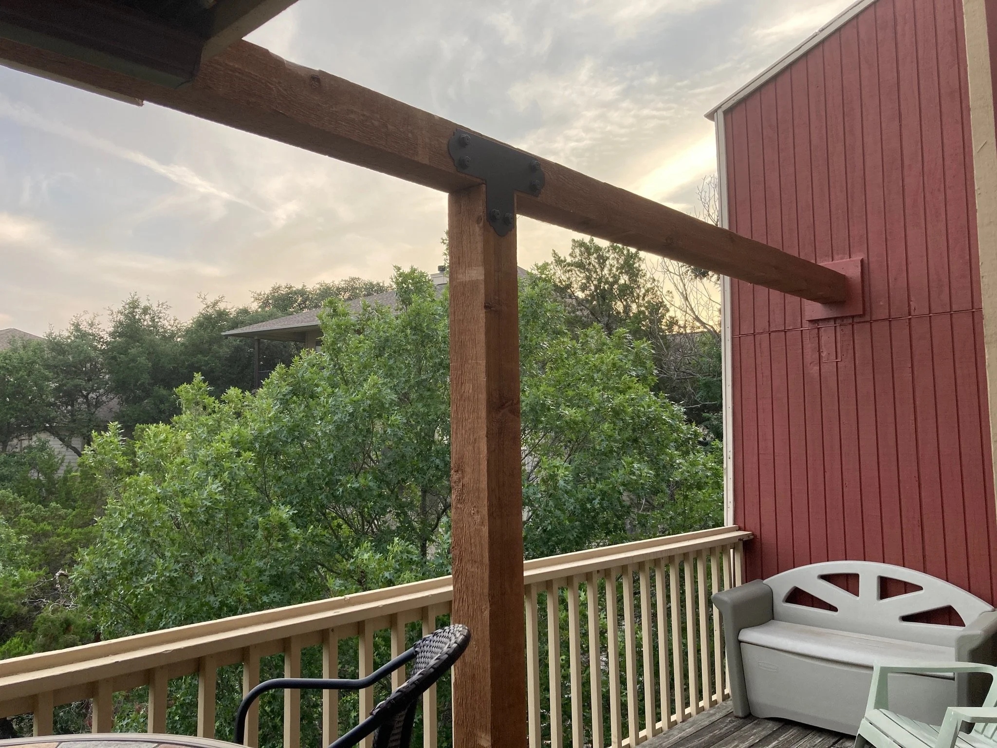 View of a wooden balcony with a white plastic chair and a black chair, overlooking green trees and neighboring houses under a cloudy sky during sunset.