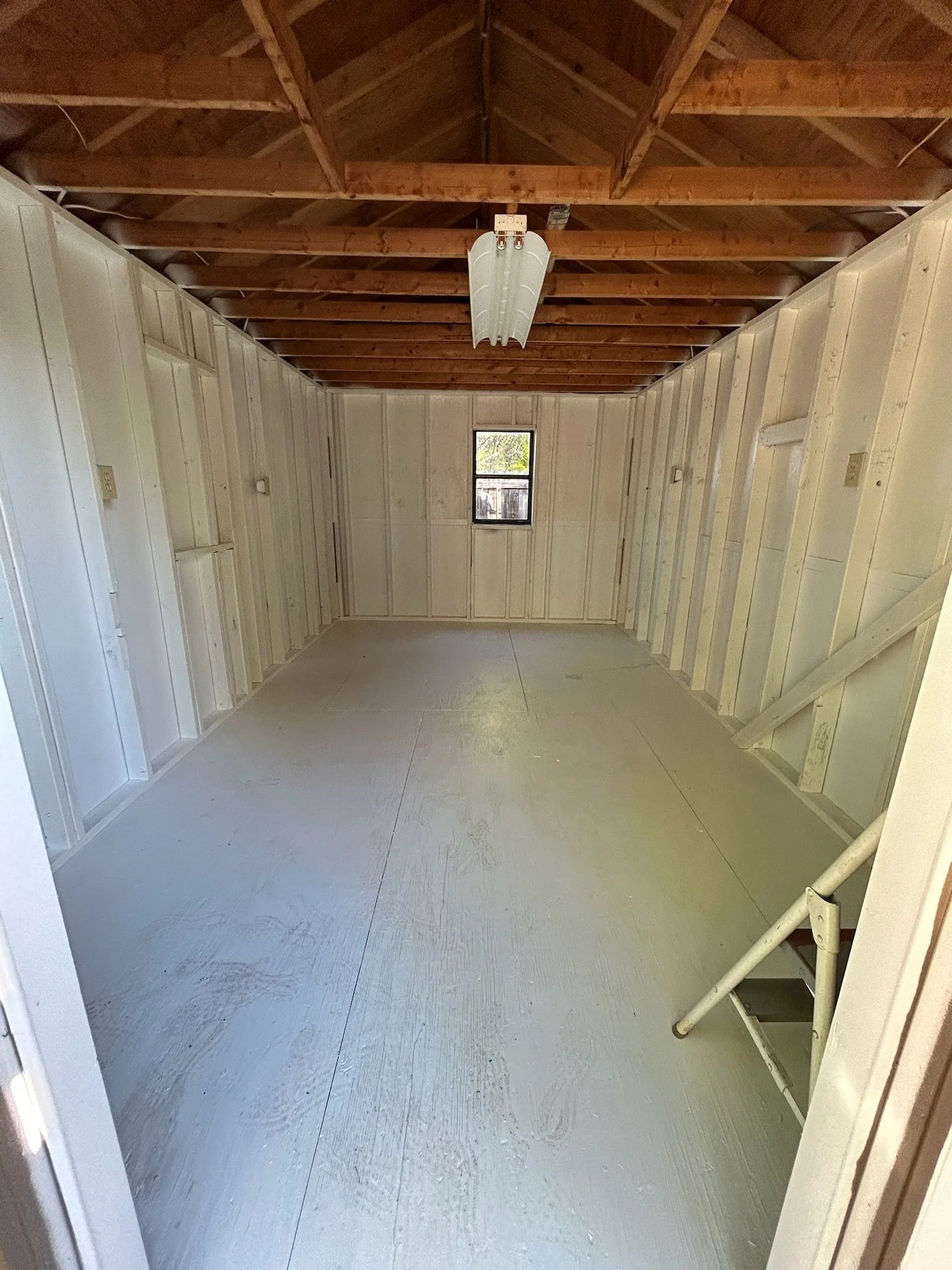 An unfinished room with exposed wooden ceiling beams, white painted walls, a small window at the far end, and a white painted floor. Part of a metal staircase railing is visible in the lower right corner.