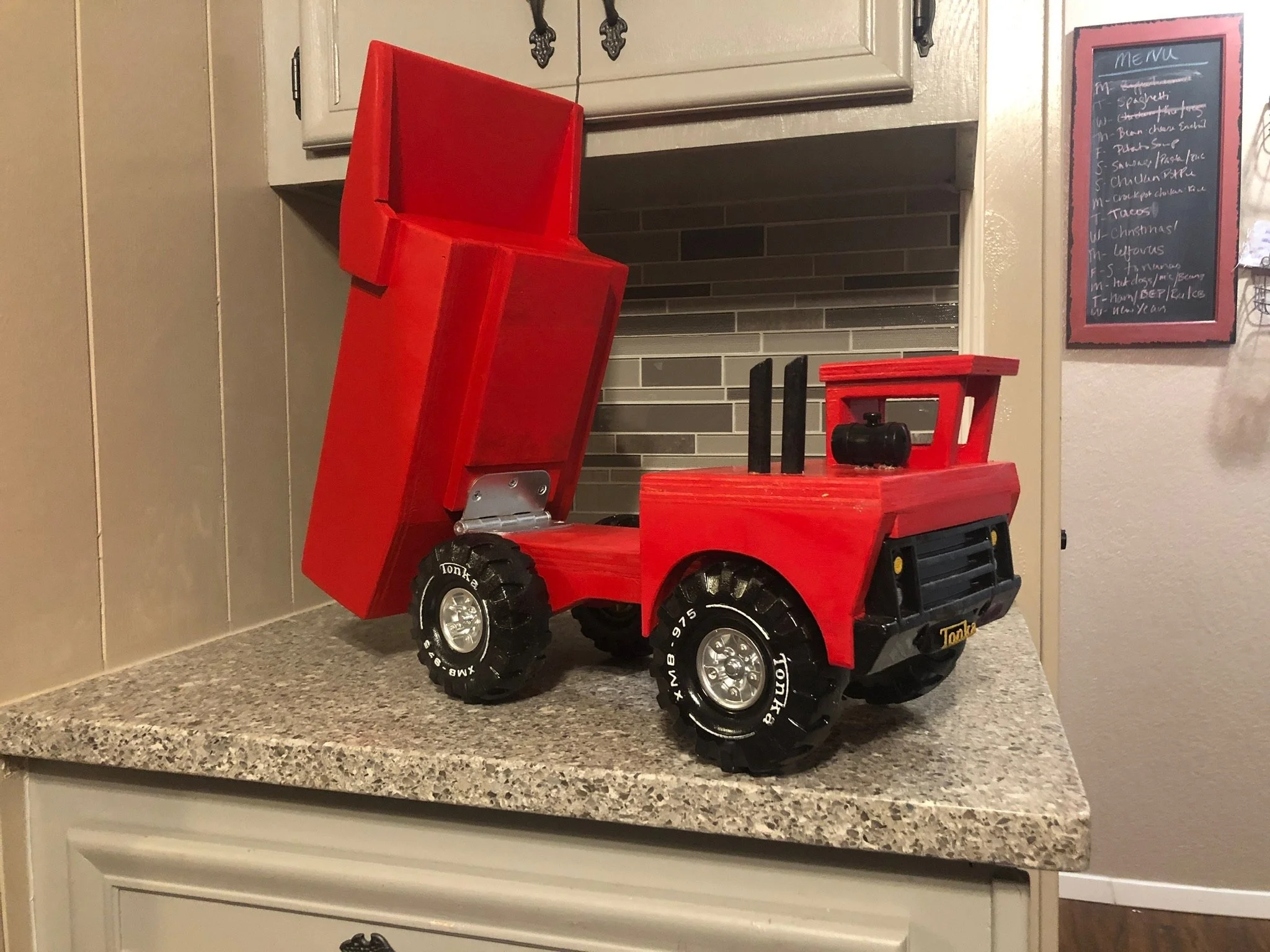 Red toy dump truck on a kitchen countertop in front of a brick fireplace and a chalkboard menu on the wall.