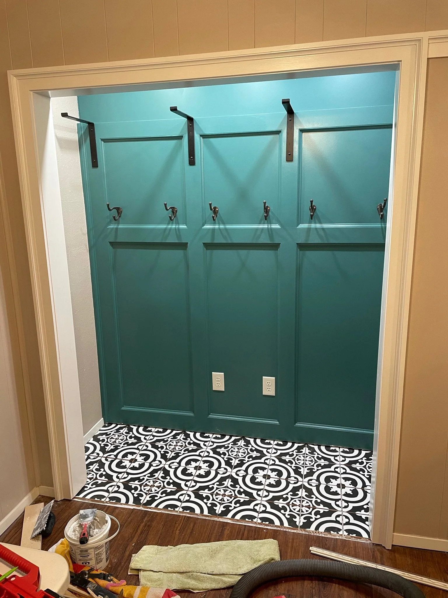 The image shows an interior space with a green painted wall and a black-and-white patterned tile floor. The wall has black hooks and support brackets, likely for hanging items. Construction or renovation tools and materials are visible on the wooden 
