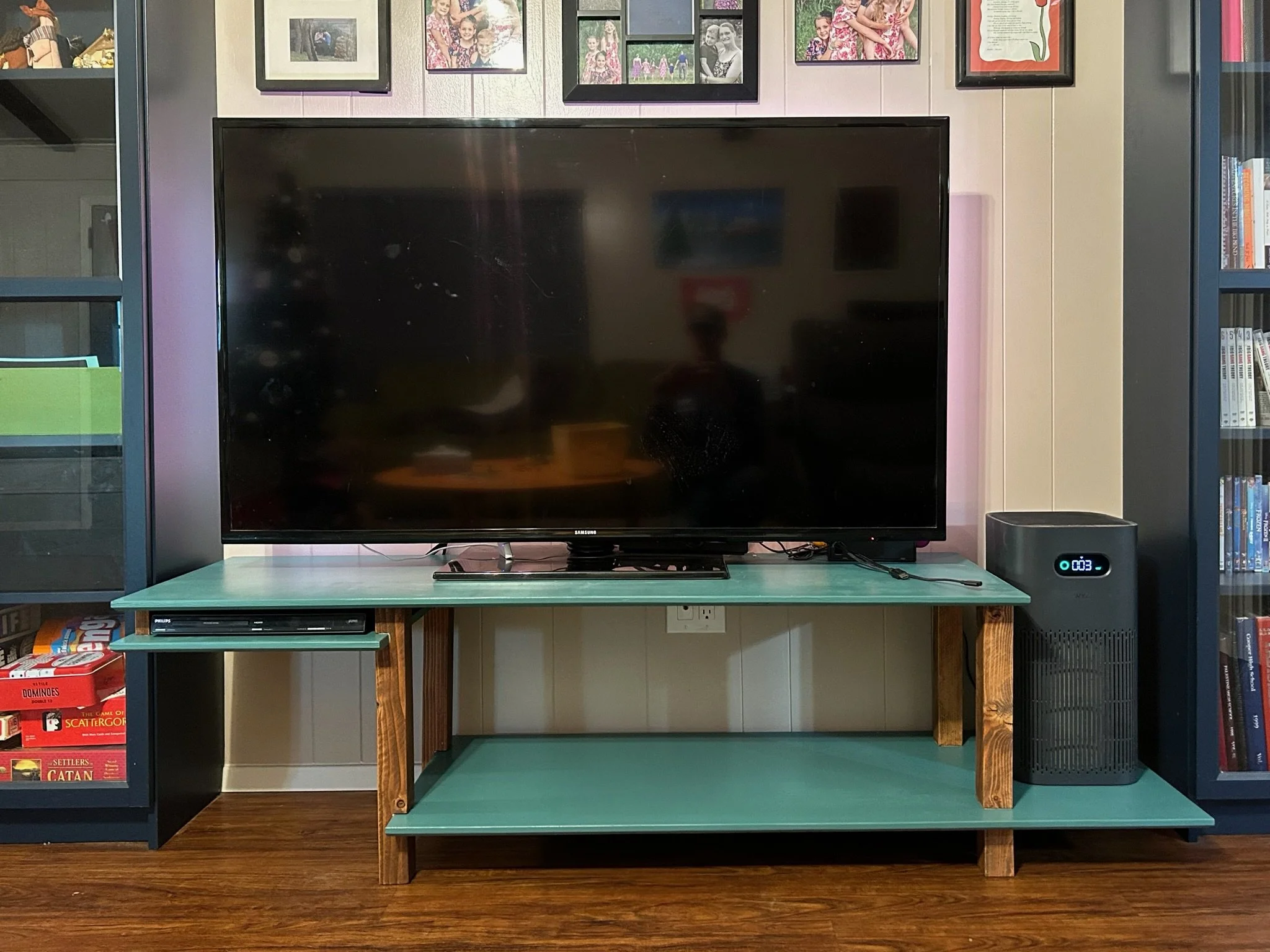 A flat-screen television on a wooden stand, surrounded by framed family photos on the wall, books on shelves, and a game box on the lower shelf.