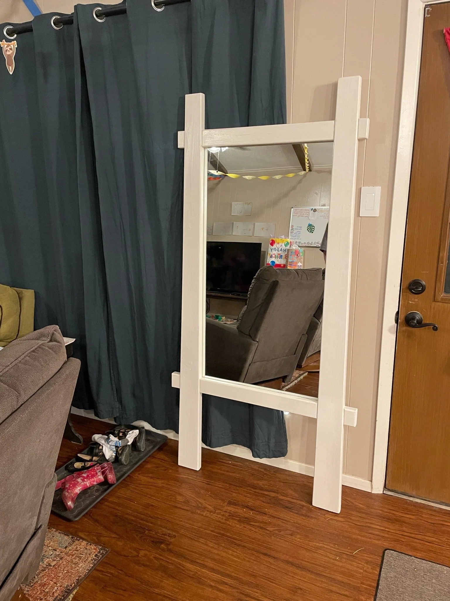 Living room with a large mirror, a couch, a shoe rack with shoes, a rocking horse, a dining table with chairs, and a doorway. The mirror reflects part of a television and children's artwork on the wall.