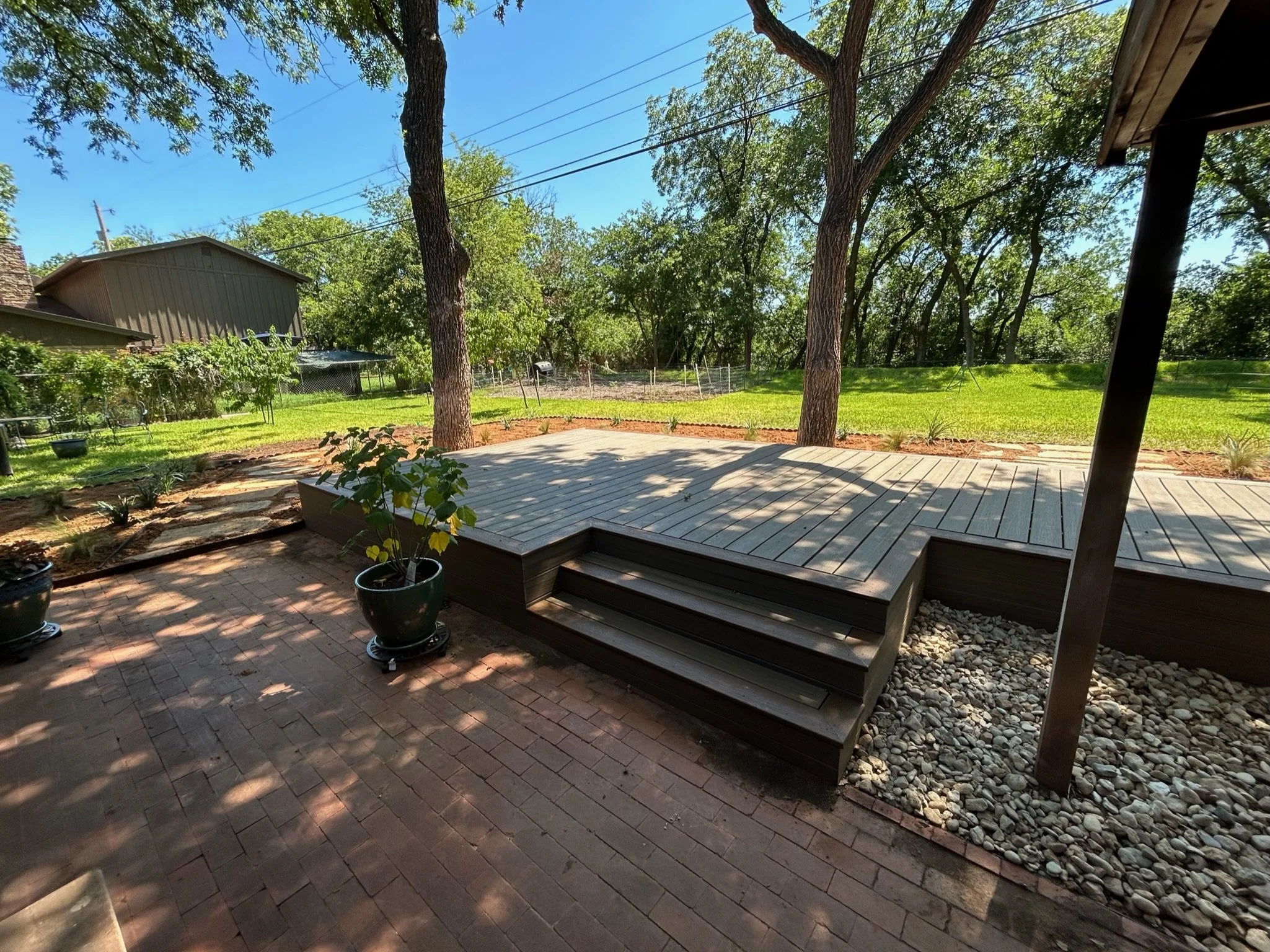 A backyard with a wooden deck, several potted plants, a garden bed with young plants, green grass, trees, and a fence on a sunny day.