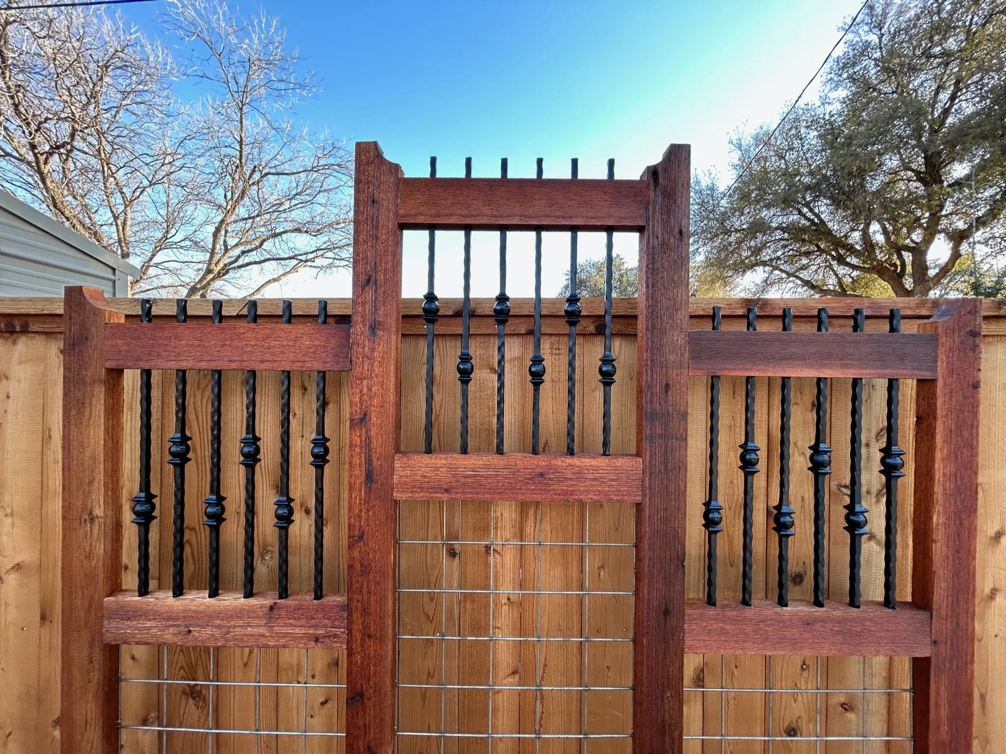 A wooden fence with decorative black metal pickets, showing a small gate with a wire latch, and a background of cloudy blue sky and leafless trees.